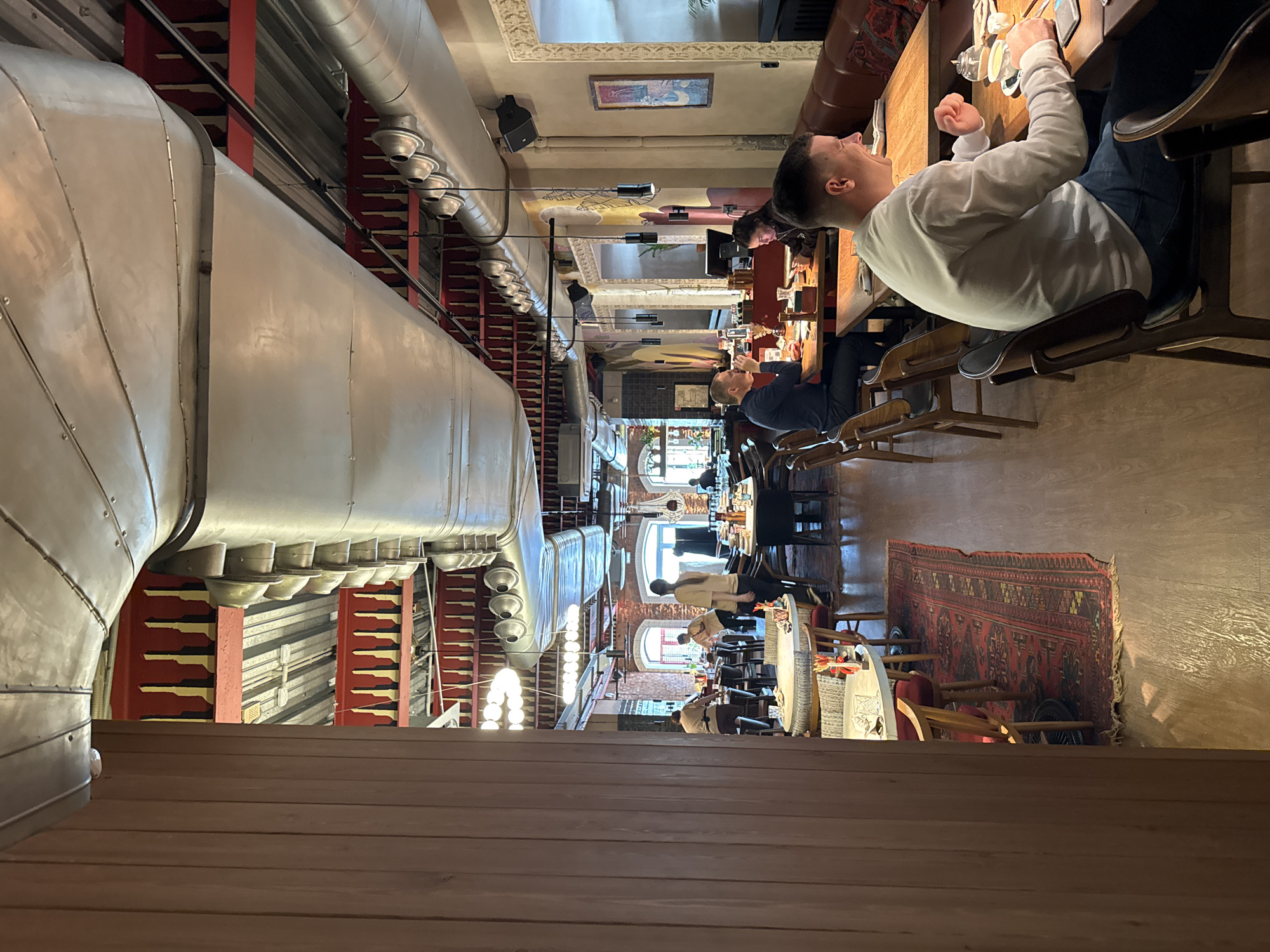 The interior of a restaurant in Izhevsk, Russia, is depicted in this vertically oriented image. The restaurant is filled with patrons seated at wooden tables and chairs along the length of the room. The décor is industrial and modern. Large silver pipes are mounted near the ceiling, with red and dark-gold colored accents on the walls. A red oriental rug rests on the floor. Patrons are engaged in various activities such as eating, talking, and using phones. The lighting is bright, with natural light coming from windows at the end of the room.