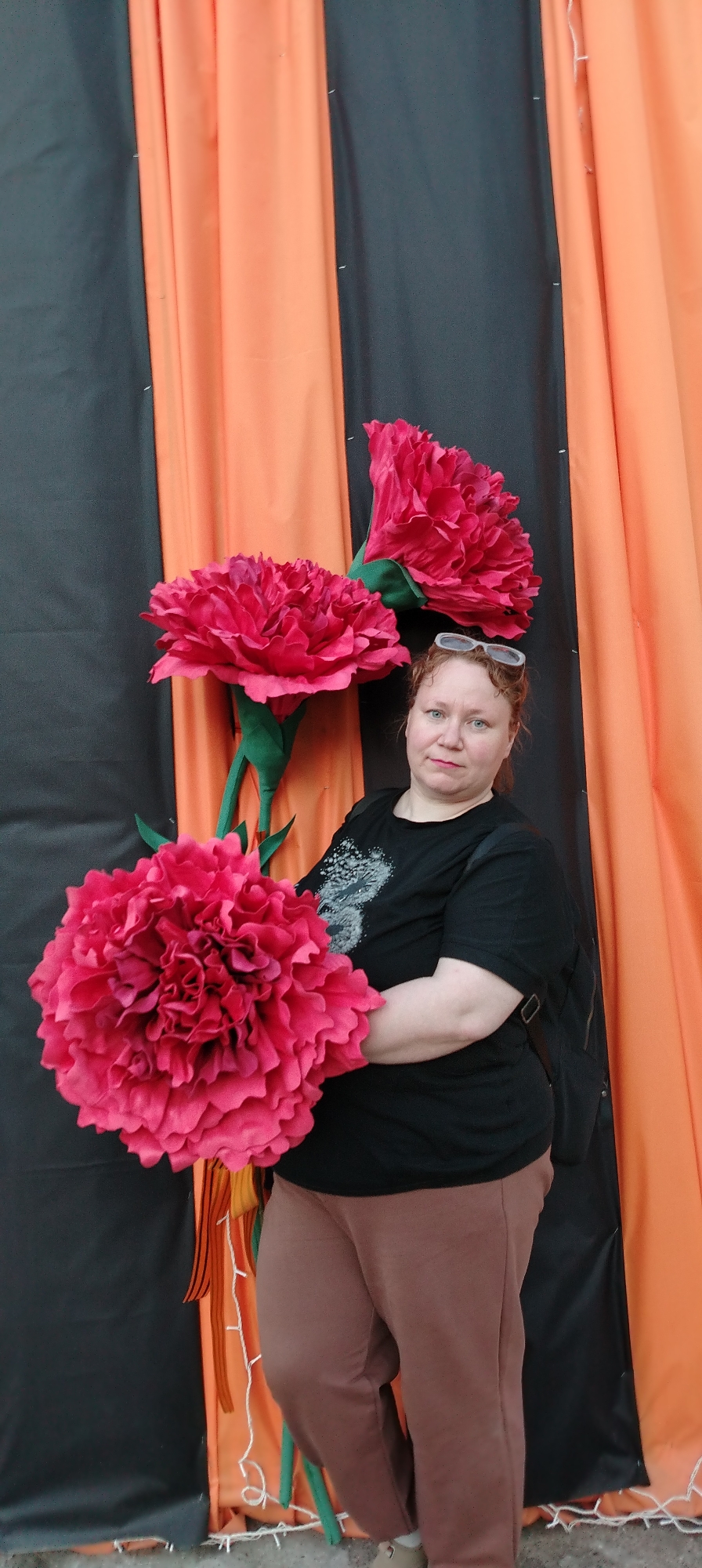 A woman stands next to three large artificial red flowers against a backdrop of alternating black and orange fabric panels. She is wearing a black t-shirt, brown pants, and has a black backpack on one shoulder. Eyeglasses are perched on top of her head. The setting appears to be outdoors in what is identified as Chaikovsky, Russia.