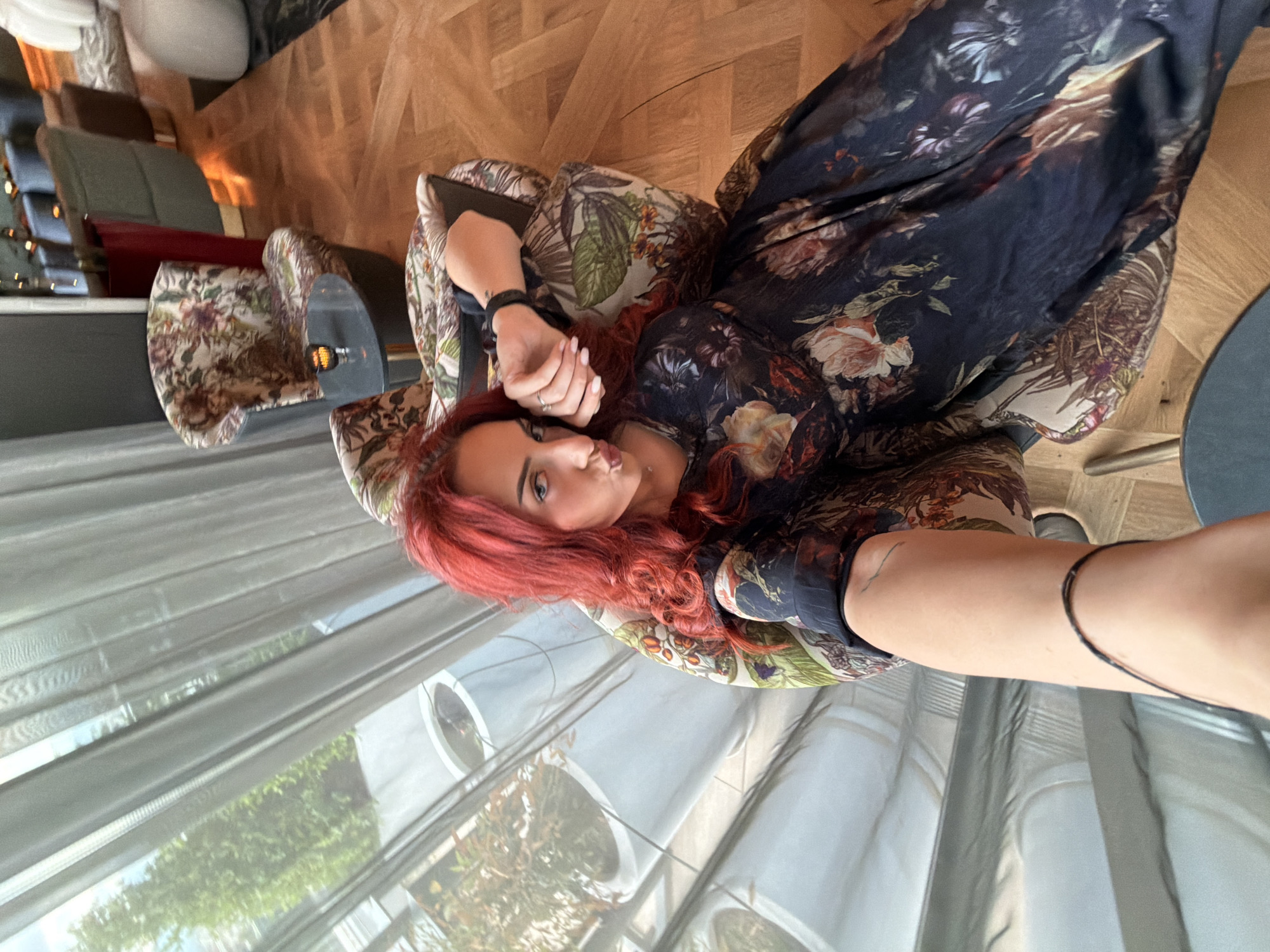 A woman with red hair is reclining in a chair with a floral pattern, wearing a long, dark floral-print dress. She is facing the camera and extending her right arm towards it. The scene appears to be indoors, possibly in a cafe or lounge area with a view of foliage through the windows, a partial view of tables, chairs, and wooden flooring. The location may be in the city of Sofia, Bulgaria.