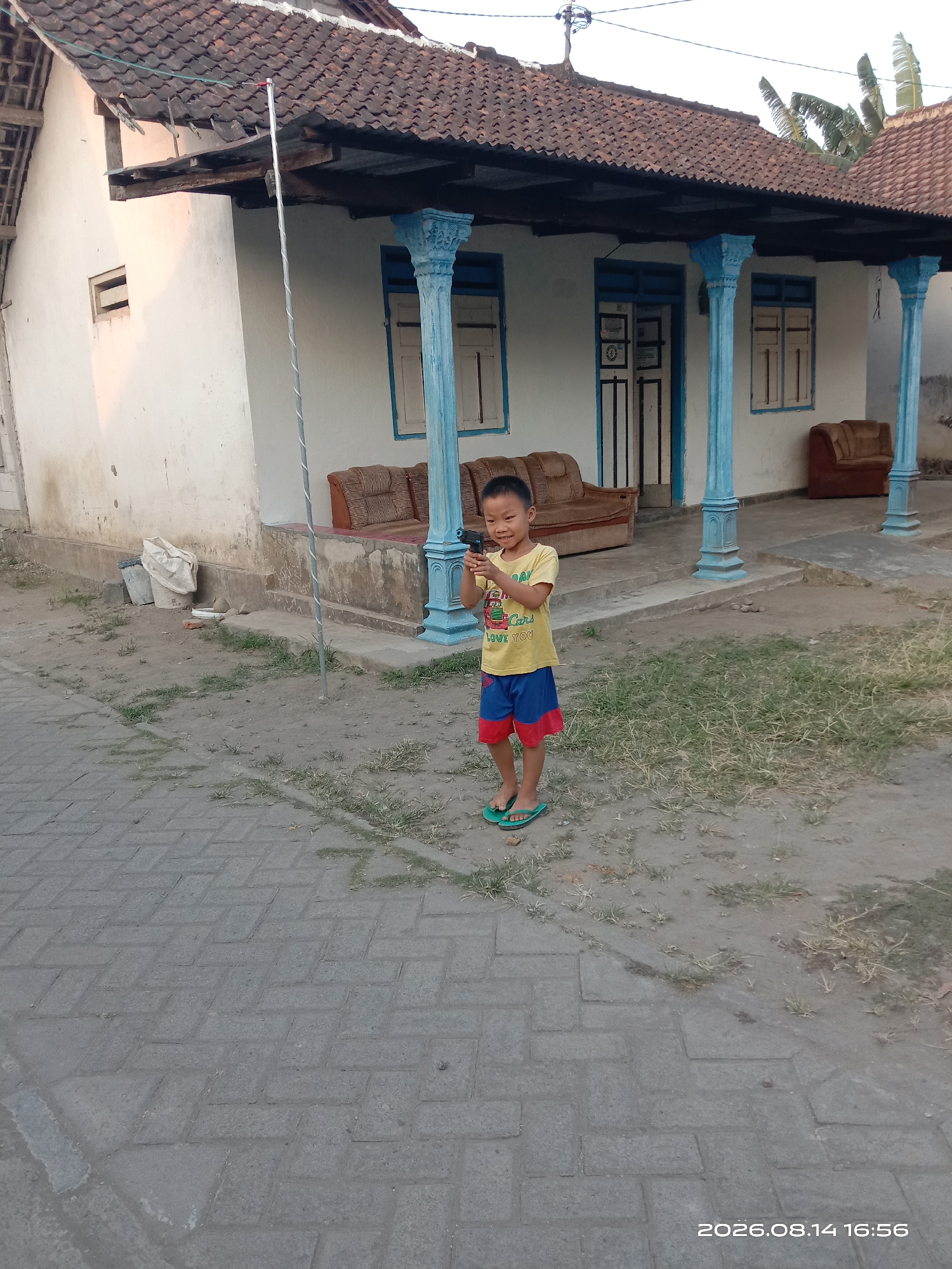 A young boy stands on a paved walkway in front of a white house with a terracotta tiled roof. He is wearing a yellow t-shirt with a graphic and the words "Cars Love You," and blue and red shorts. He is holding a black object that resembles a toy gun. The house has blue-trimmed windows and decorative blue pillars supporting an overhang. Brown upholstered furniture is visible on the porch. The ground in front of the house is a mix of dirt and sparse grass. A trash can is visible to the left of the house. The image is dated 2026.08.14 16:56. The location is identified as Banyuarang, Indonesia.