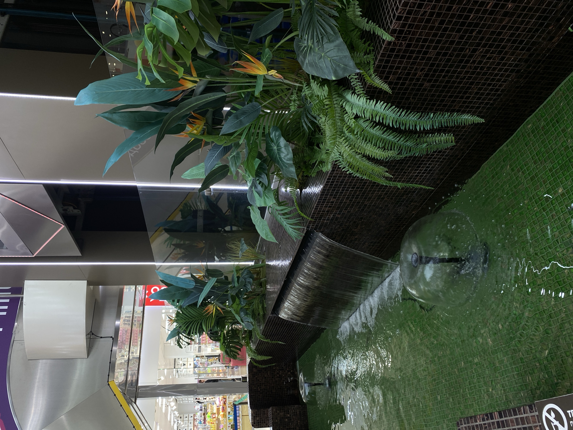 The image depicts an indoor water feature in a commercial setting, possibly a shopping mall in Sochi, Russia. The water feature consists of a cascading waterfall effect over a green tiled surface. A spherical fountain component is partially submerged, creating ripples in the water. Lush greenery, including various types of ferns and flowering plants with orange and yellow blossoms, is integrated into the structure. The background shows glimpses of a shopping area, with shelves stocked with products and shoppers moving about. Overhead lighting fixtures illuminate the scene. The composition suggests the fountain is intended to create a relaxing oasis within the shopping environment.