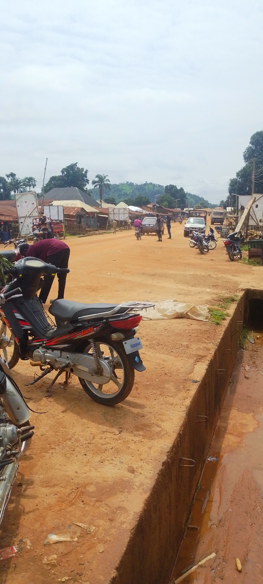 The image depicts a street scene in Baissa, Nigeria, characterized by a dirt road and structures on either side. In the foreground, several motorcycles are parked, with one prominently featured on the left side of the frame, facing towards the viewer. A person wearing a dark red shirt is bent over near this motorcycle, possibly attending to it. Further down the street, other vehicles, including cars and motorcycles, are visible. People are scattered throughout the scene, some walking, others riding motorcycles or standing. The buildings lining the street appear to be a mix of shops and residences, with corrugated metal roofs being a common feature. The sky is overcast. A concrete drainage channel runs along the right side of the road, containing murky water and some debris. The overall impression is that of a busy, everyday scene in a Nigerian town.