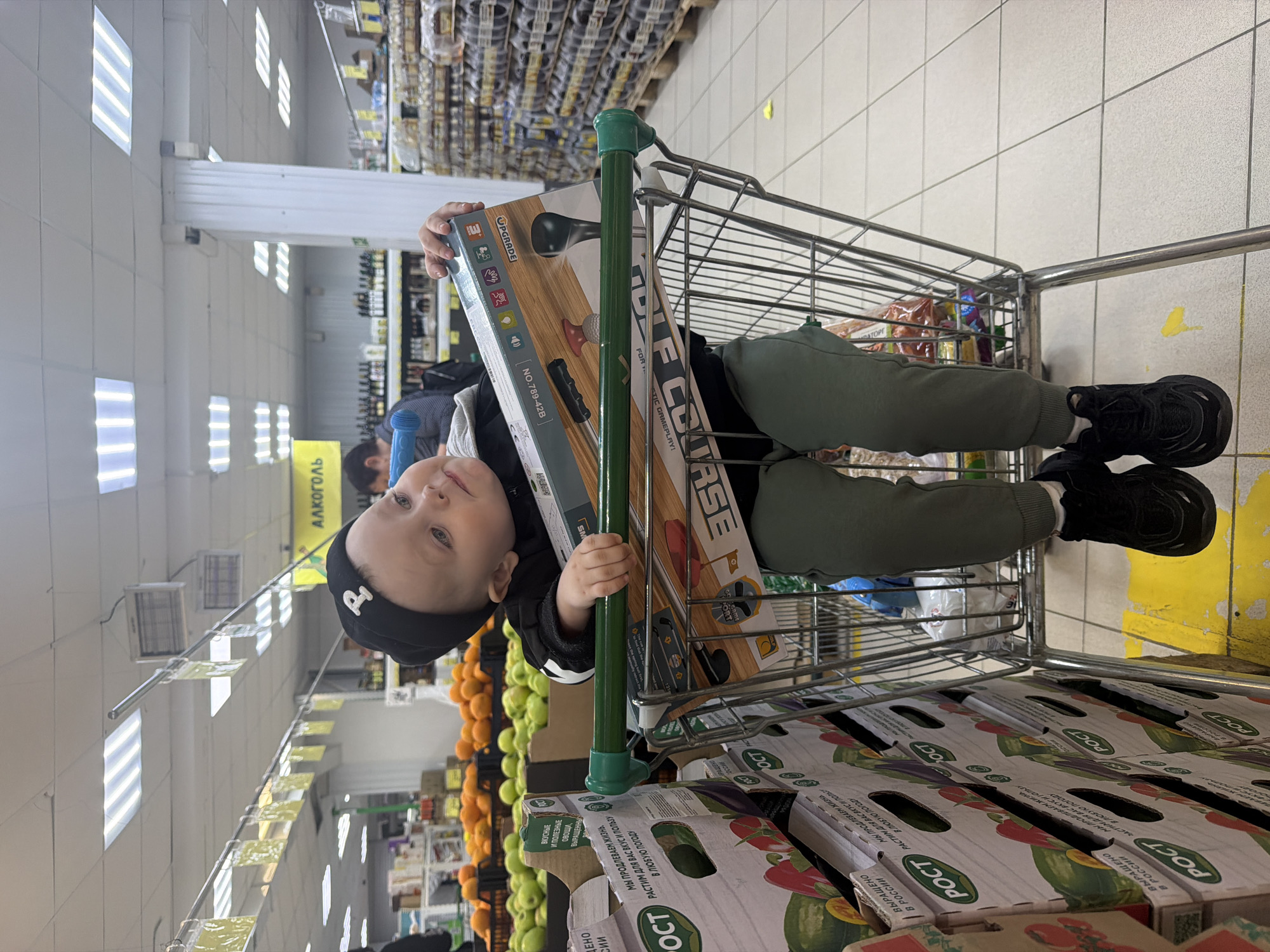 The image shows a young child lying in a shopping cart at a grocery store. The child is wearing a black beanie, a gray jacket, and green pants, and black shoes. The child is holding a boxed toy game titled "Upgrade". The shopping cart is filled with boxes that appear to be vegetables. A row of oranges and other fruits are visible in the background, as well as a sign that reads "ALKOHOL." The location is identified based on the OCR data as somewhere in Russia, specifically where the ROST vegetables are produced. The location is more specifically inferred as a grocery store in Chaikovsky, Russia.