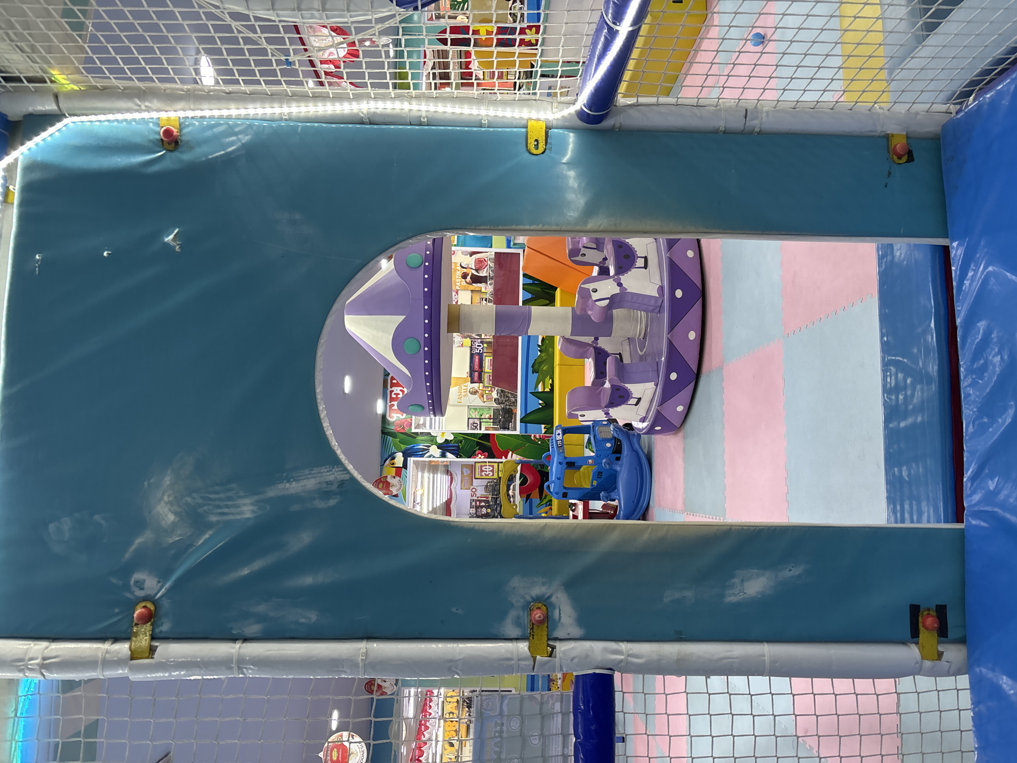 The image depicts the interior of an indoor playground. A blue, soft-edged archway frames a view of a purple and white carousel with horse-shaped seats. To the left of the archway, a blue rocking toy resembling a truck is visible. The floor is covered with interlocking foam mats in pink and light blue. In the background, there are signs and displays suggesting a commercial setting, possibly a mall or a store. The walls and structures of the playground are padded with blue and white material, and white netting encloses some areas. The scene is brightly lit with artificial lighting. The image is taken from within the playground, looking through the archway into the play area. No people are visible in the frame.
