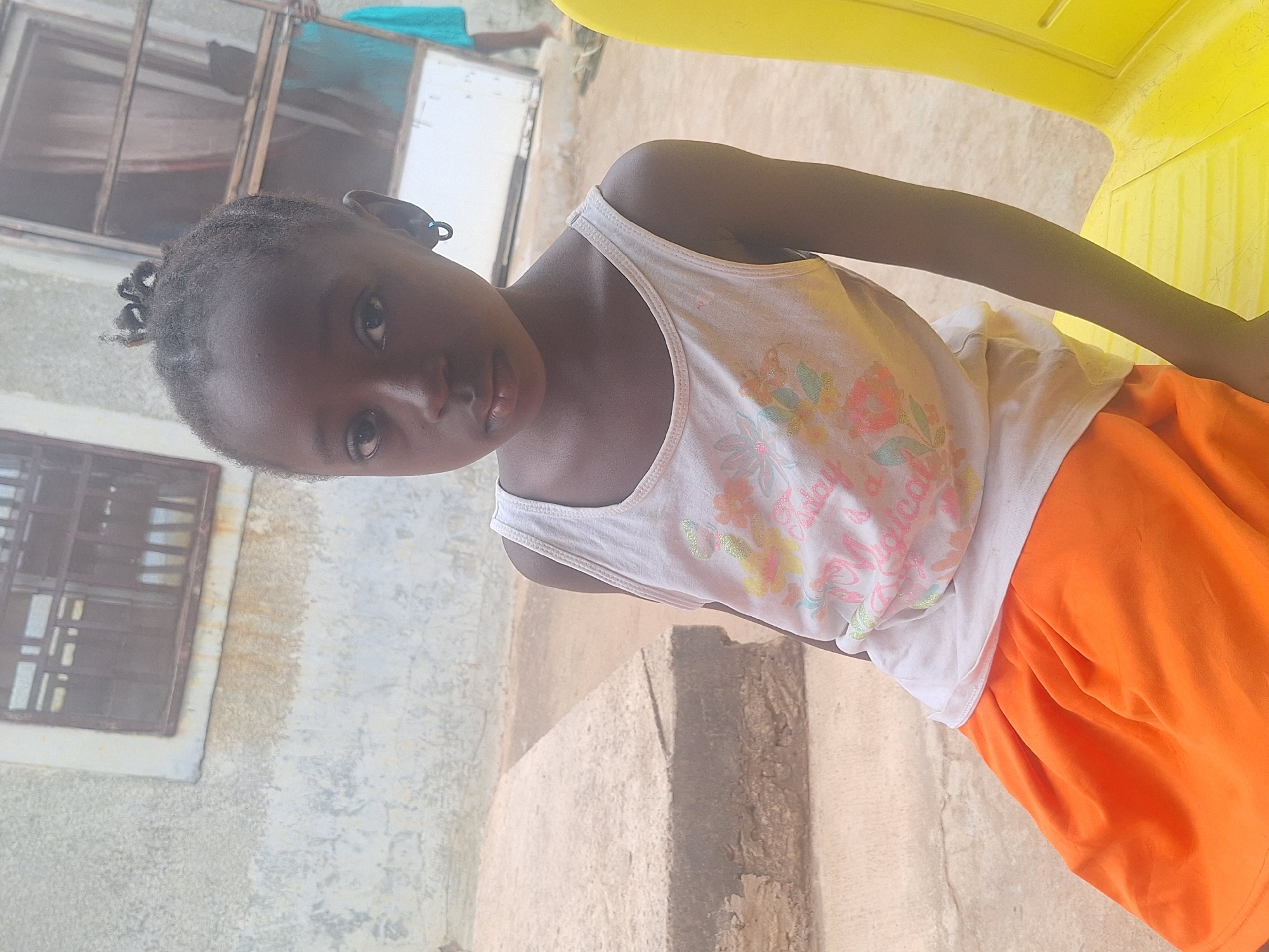 A young Black girl in Sabon Tasha, Nigeria, stands outdoors. She wears a white tank top with floral designs and the word "Magical" in pink letters. She is wearing an orange skirt. Her hair is braided in small buns. She appears to be leaning against a yellow plastic chair. In the background are sections of a weathered, white-washed building with a window. A concrete stairway is also in view.
