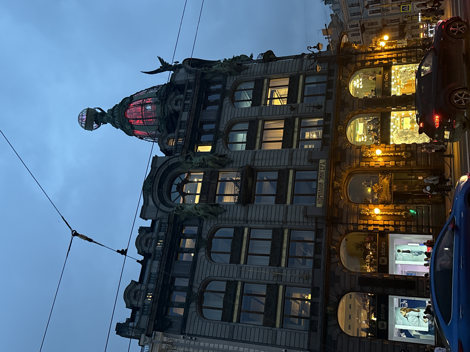 The image shows the Singer House, also known as the Dom Knigi (House of Books), located on Nevsky Prospekt in Saint Petersburg, Russia. The building features a striking Art Nouveau architectural style with a prominent glass dome topped by a globe sculpture. The sky is a deep blue, indicating it is dusk or early evening.
The facade of the building is ornate, with multiple stories of arched windows. Some windows are illuminated from within, showing glimpses of interiors that appear to be shops and cafes. The ground floor features large display windows advertising "VK store" and "Cafe René." People are visible inside these establishments, as well as on the street.
In the foreground, two cars are driving on the street. A blue car is on the left, and a dark SUV with its brake lights illuminated is on the right. Wires for an overhead trolley system are visible crisscrossing the sky. Streetlights and architectural lighting cast a warm glow on the scene. The overall impression is of a lively urban environment at twilight.