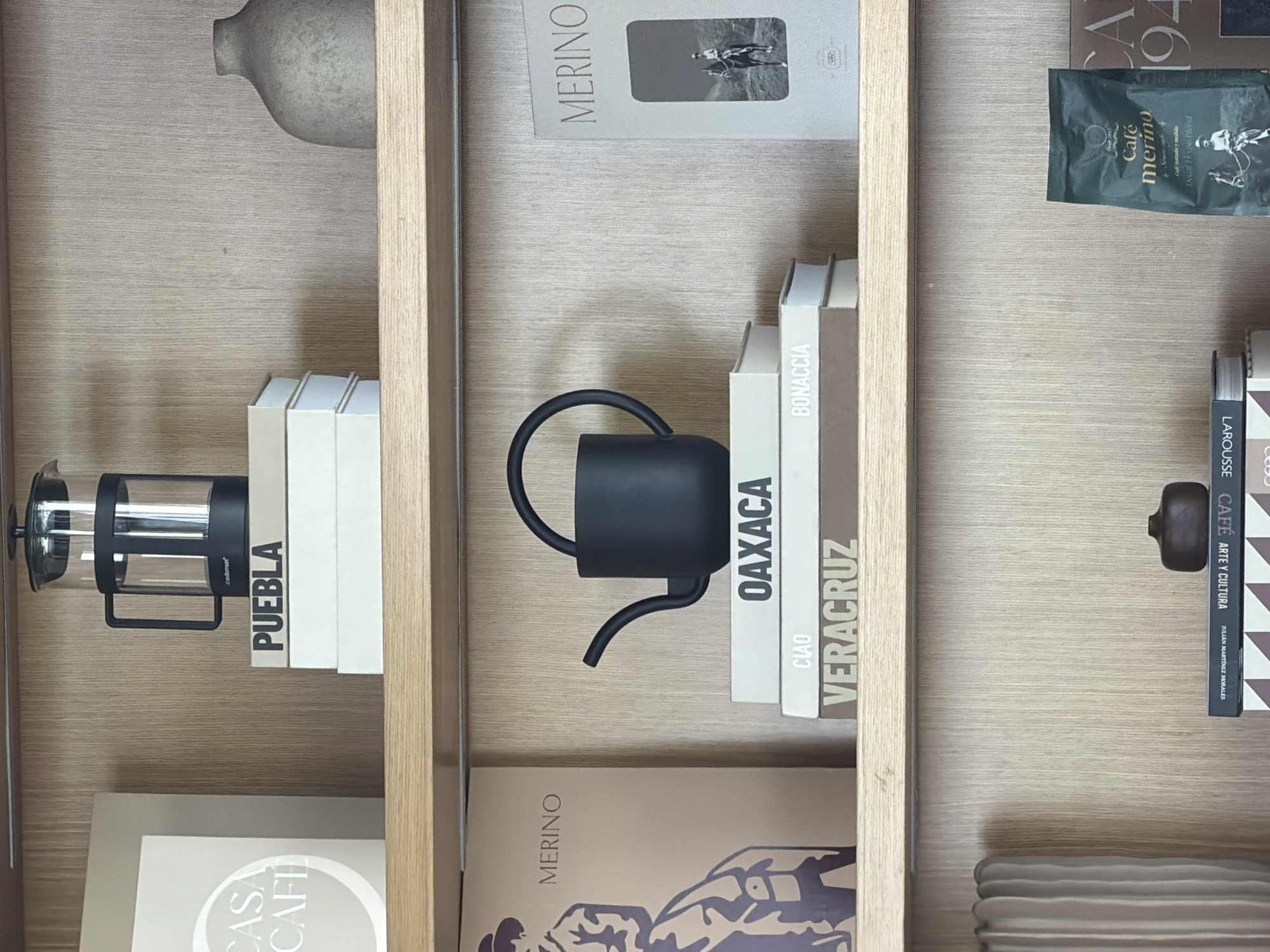 A shelf contains various objects related to coffee. On the left shelf, a French press coffee maker sits next to books labeled "PUEBLA" and "CASA CAFE". The middle shelf contains a black coffee kettle and books labeled "OAXACA", "CIAO", "BONACCIA", and "VERACRUZ", along with a book labeled "MERINO". On the right shelf, a bag of "Café merino" coffee is next to books labeled "LAROUSSE CAFE ARTE Y CULTURA", and a "CA 194" book. A small dark-colored object also sits on this shelf. The shelves are made of light-colored wood. The scene appears to be indoors, likely in a home or a shop related to coffee. San Bernardino Tlaxcalancingo is not discernible from the image.