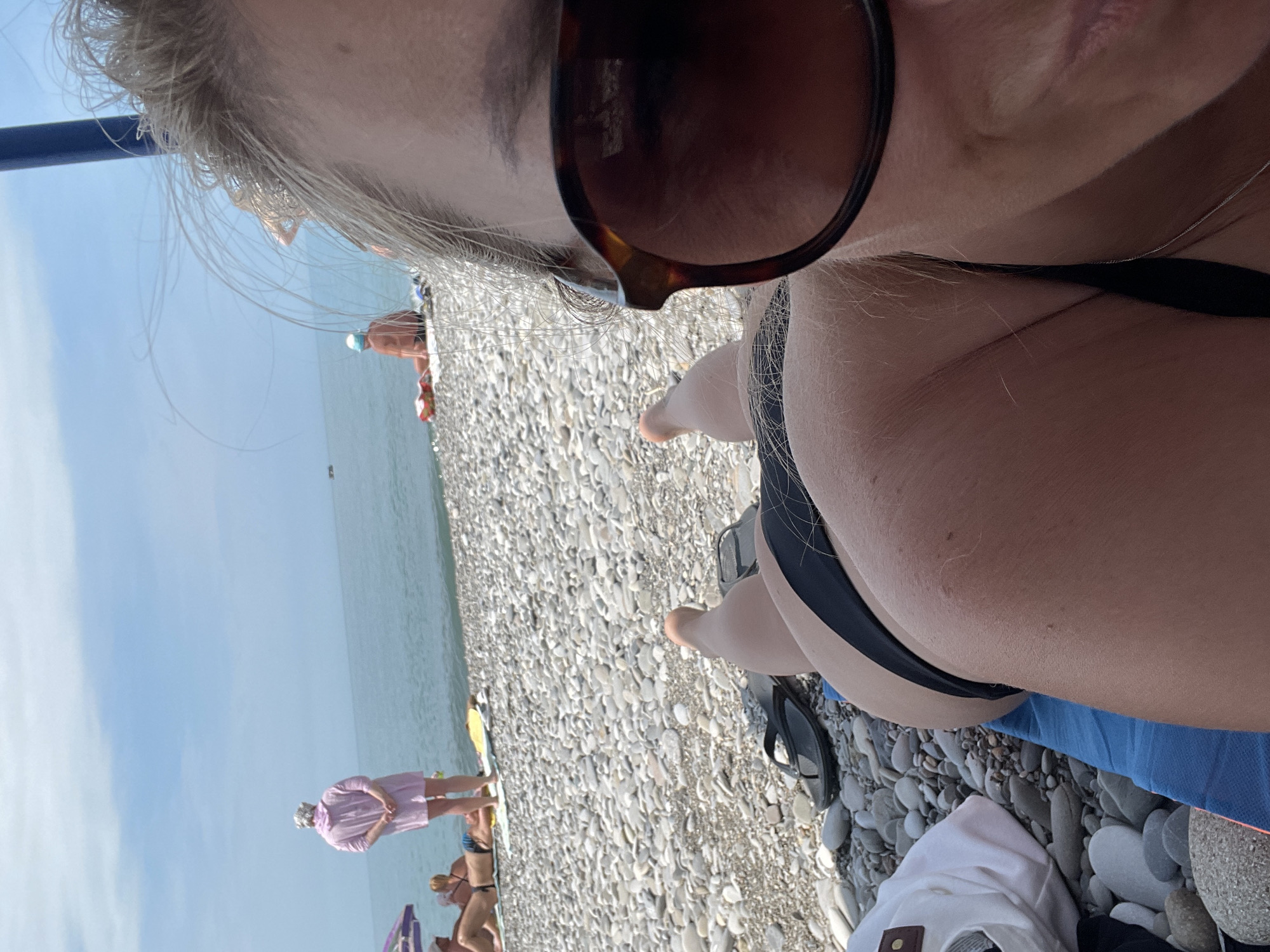A woman lies on a blue towel on a pebbly beach, facing away from the camera and looking towards the sea. She is wearing a black bikini. Her face is visible in the foreground, partially obscured by large, dark sunglasses. In the background, other people are visible on the beach and in the shallow water. The sky is a pale blue with some clouds. The location is the beach in an unspecified city.