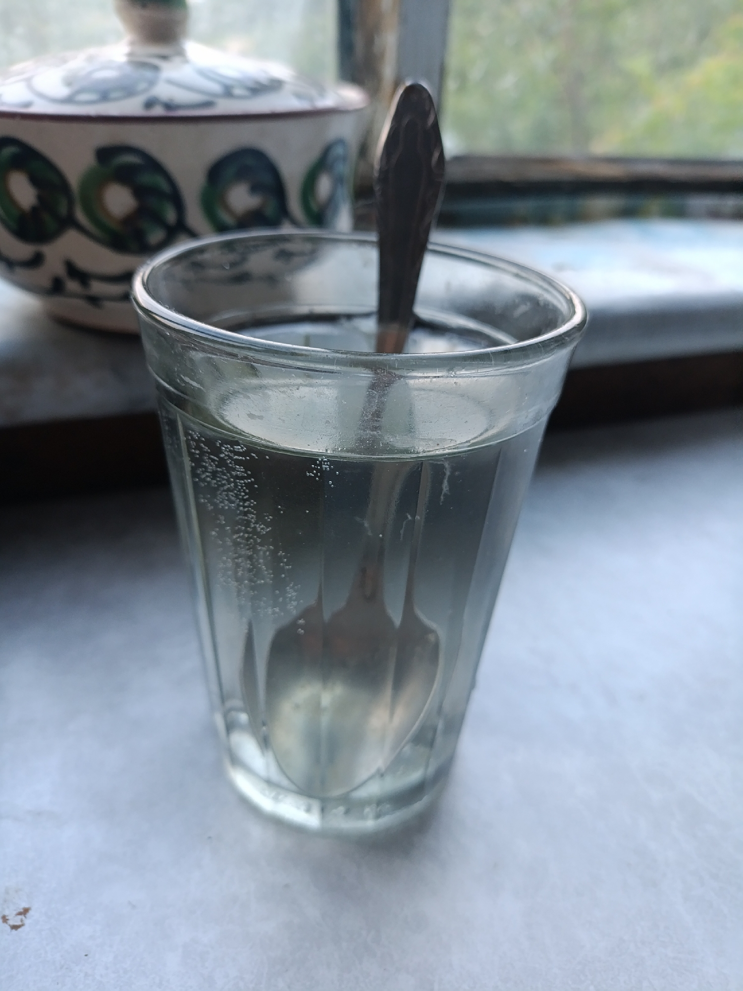 An overhead shot captures a glass of clear liquid with a spoon submerged in it, resting on a textured, light-colored surface. Bubbles are visible rising within the liquid, suggesting it is carbonated. In the background, a blurred, decorative bowl with a lid, featuring a white base with green and blue circular patterns, sits near a window. The window frames a slightly out-of-focus view of greenery and a blurred ledge. The scene is well-lit, possibly by natural light from the window. The location is identified as Koklaisai, Kazakhstan.
