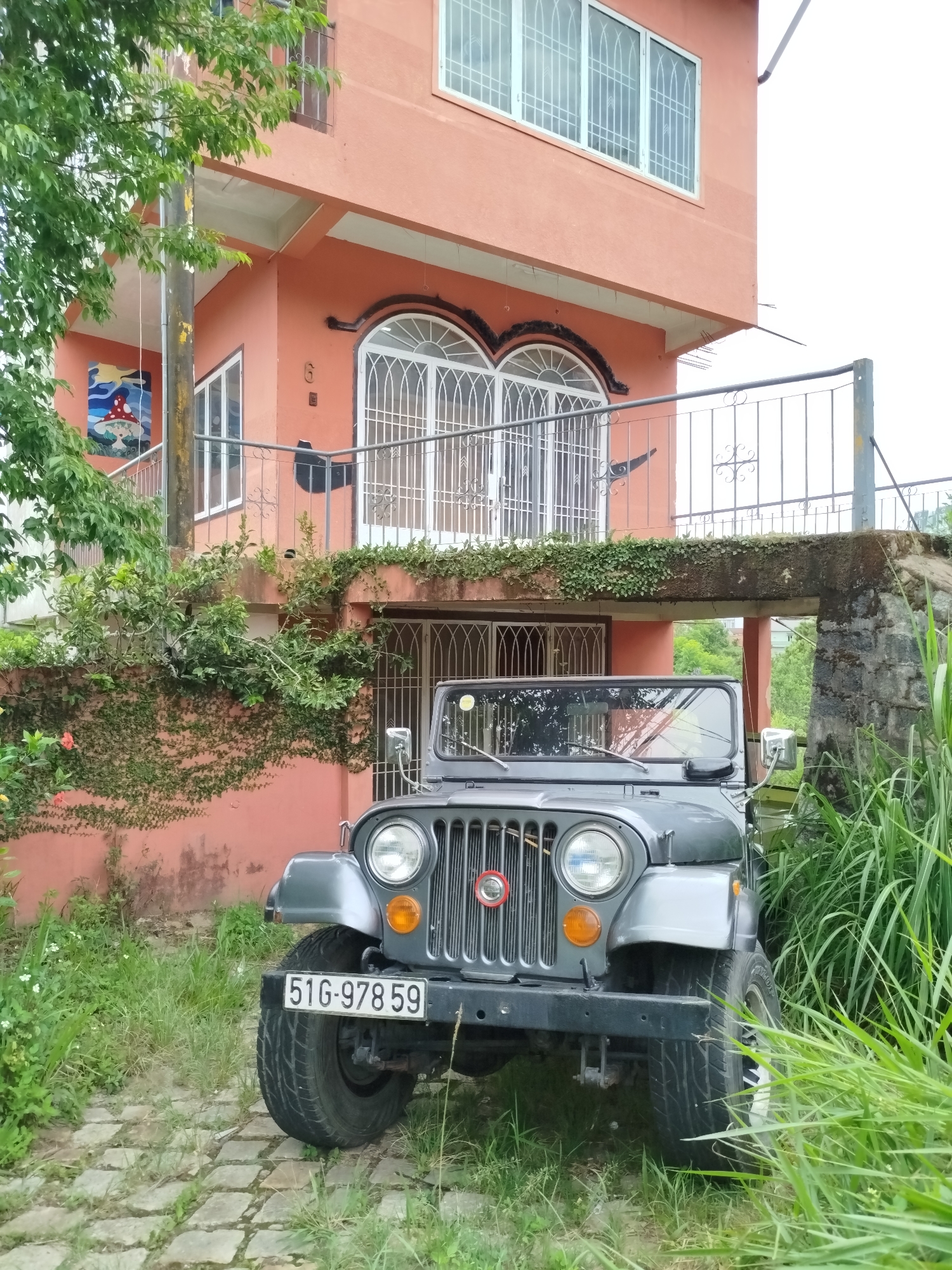 A grey Jeep Wrangler stands parked in front of a salmon-colored two-story building with metal-barred windows. The Jeep's license plate reads "51G-97859." The building features a small balcony with a metal railing above the Jeep's parking area. Green vegetation covers sections of the building's front. The ground is cobbled with stone pavers. Tall grass grows on the right side of the Jeep. The setting appears to be an urban area, possibly Da Lat, Vietnam. A mushroom-themed artwork hangs on the building's exterior wall.