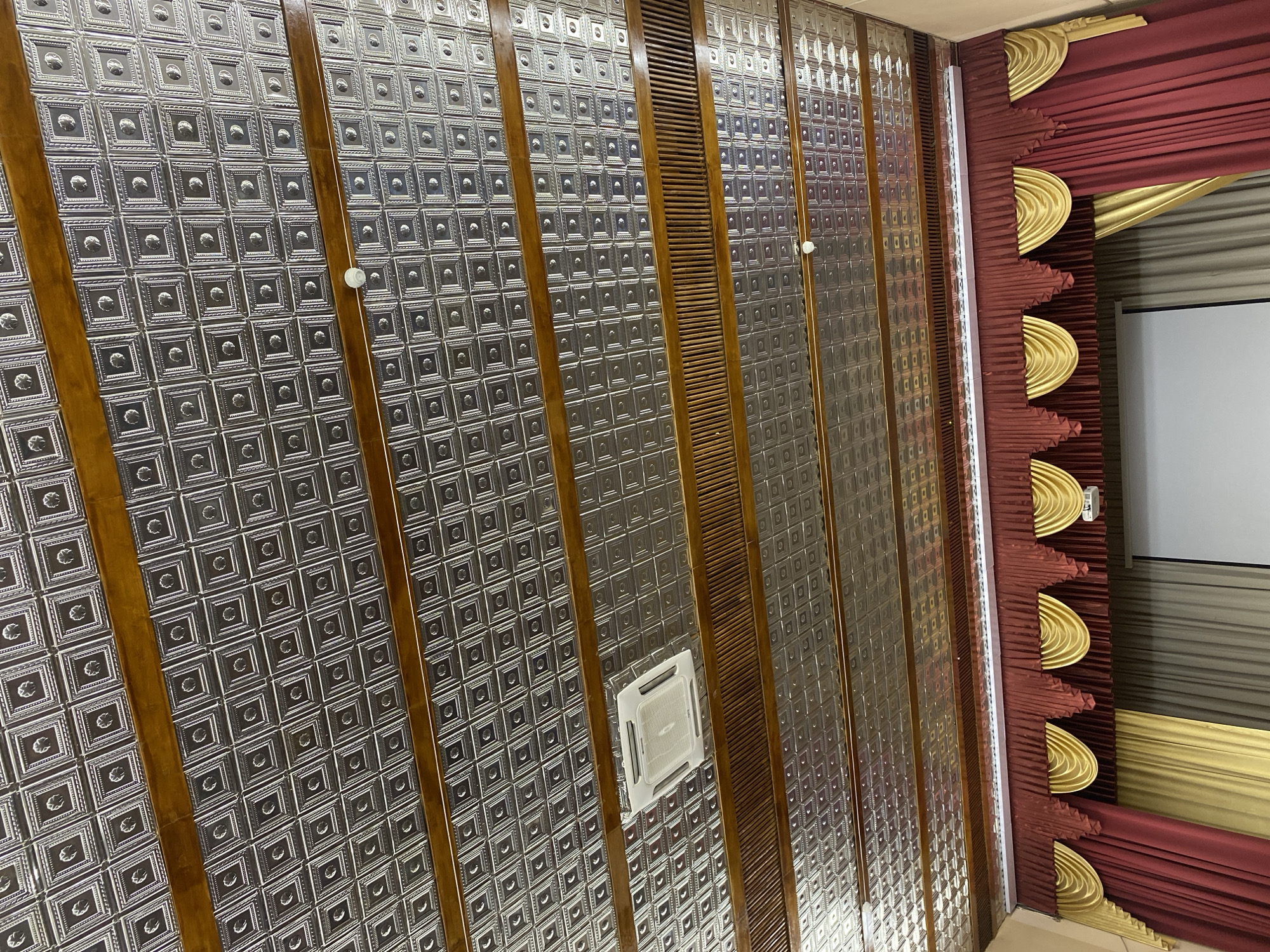 The image captures an ornate indoor ceiling and an adjacent wall, viewed from a low, upward-angled perspective. The ceiling is predominantly covered with silver-colored, embossed metal tiles, each featuring a raised square frame surrounding a central circular pattern. These decorative panels are separated by parallel, polished dark wooden beams or slats that span the length of the ceiling, creating a structured, striped appearance. One of these wooden sections also includes a slatted vent. A white, square cassette-type air conditioning unit is integrated flush into the ceiling. Small, circular white light fixtures are visible within the ceiling panels, and a long, linear LED light strip illuminates the edge where the ceiling meets the wall on the right.
The right side of the frame reveals a dramatic wall treatment featuring rich burgundy and gold drapery. The burgundy fabric is intricately pleated and adorned with prominent, layered gold, fan-shaped decorative elements, creating a theatrical aesthetic. A large white projection screen is partially visible beyond the drapery, suggesting the setting is likely an auditorium, cinema, or a grand performance/presentation hall in Votkinsk, Russia. There are no discernible words or legible text present in the image.