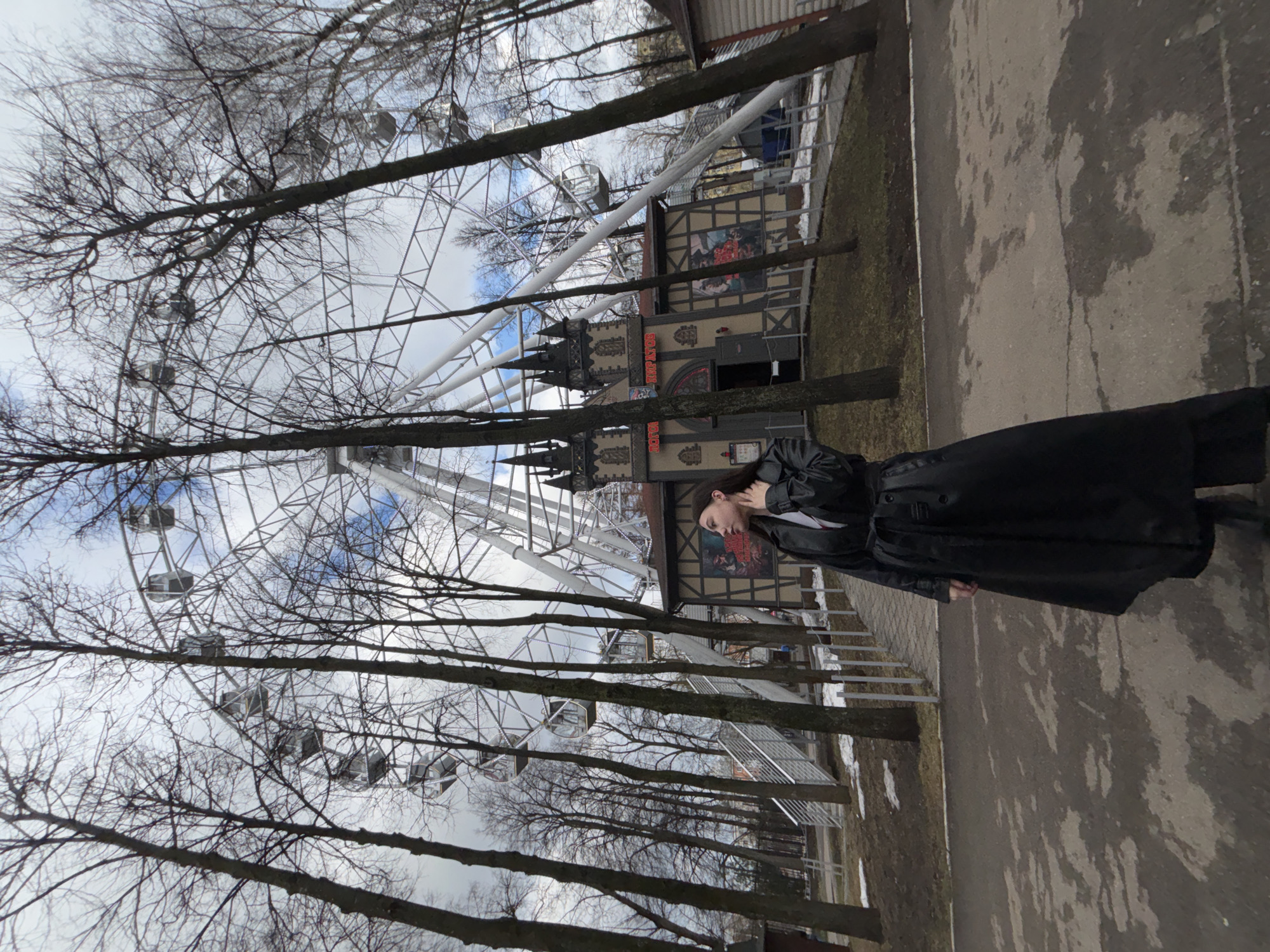 The image shows a Ferris wheel in a park in Perm, Russia. A woman in a long black coat stands to the right of the frame. Bare trees are prevalent throughout the park, partially obscuring the Ferris wheel. A building with gothic-style turrets is partially visible behind the woman and Ferris wheel. A paved pathway runs along the right side of the image. Patches of snow are visible on the ground.