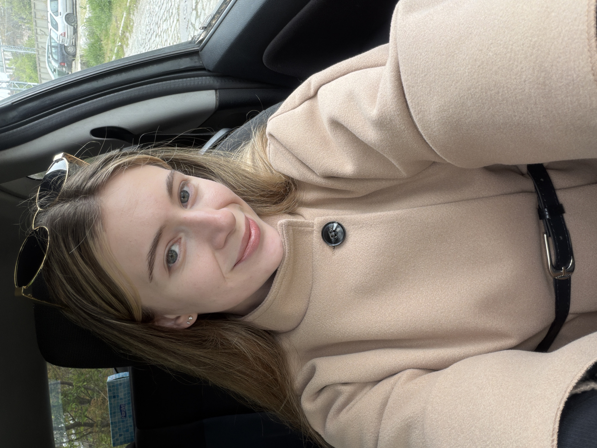 A young woman is taking a selfie in what appears to be the passenger seat of a car. She is wearing a light brown coat with a black belt. Sunglasses are perched on her head. The background visible through the car window shows a cityscape, possibly in Plovdiv, Bulgaria. Visible elements include a cobblestone street, other parked cars, and greenery. The lighting suggests it is daytime.
