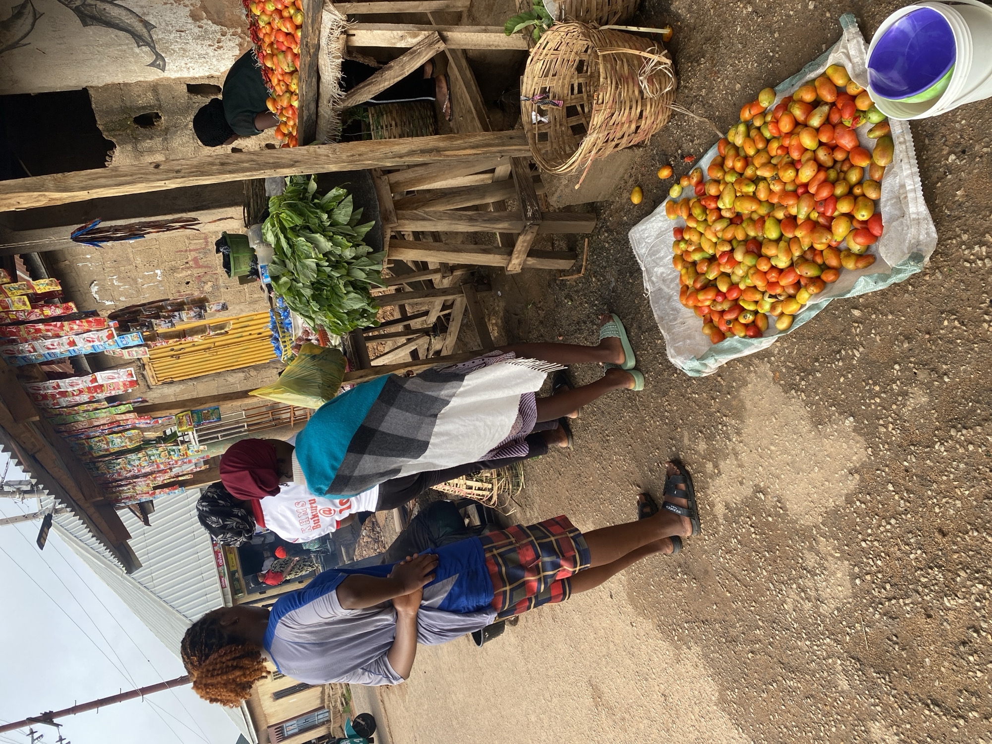 The image depicts an outdoor market scene in Jos, Nigeria. In the foreground, a pile of ripe tomatoes of various sizes and colors, ranging from red to yellow-green, is spread out on a white tarp. To the right of the tomatoes, a white bucket contains two stacked bowls, one blue and one green. In the background, several people are visible. Three individuals are standing, facing away from the camera, near a market stall. One person, wearing a blue and grey shirt and plaid shorts, stands in profile, with their hands behind their back. Next to them, another person is draped in a blue and white patterned cloth. A third person, wearing a white shirt with text that appears to read "KAMS Bukuru," stands with their back to the camera. The market stall itself is characterized by a rustic wooden structure with shelves displaying various goods. Bags of what appear to be snacks or packaged food items hang from the stall's roof. Further back, a person in a dark shirt is seated behind a display of tomatoes and leafy green vegetables. The ground is covered with gravel and dirt, suggesting an unpaved market area. The overall impression is of a busy, local marketplace in Jos.