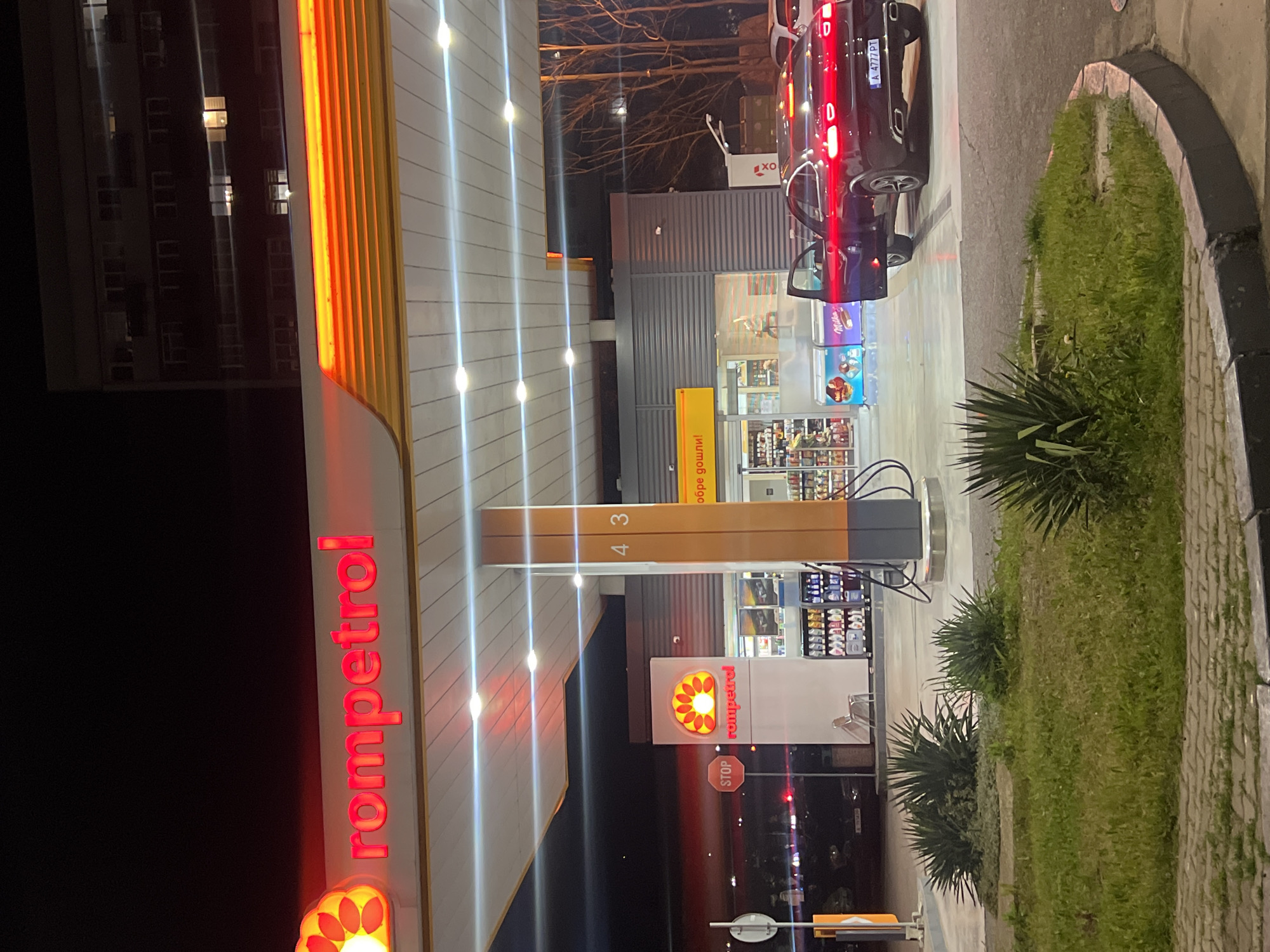 The image shows a Rompetrol gas station at night. The Rompetrol logo is prominently displayed in red neon. A dark-colored car with the license plate "A 4777 PT" is parked at one of the pumps with the driver-side door open. The gas station's convenience store is visible, with illuminated shelves of products. There is also an "XO" logo on the store. The gas pumps are numbered "4 3". A building with multiple windows is in the background. Vegetation borders the edge of the gas station area. This is Rompetrol gas station in Burgas, Bulgaria.