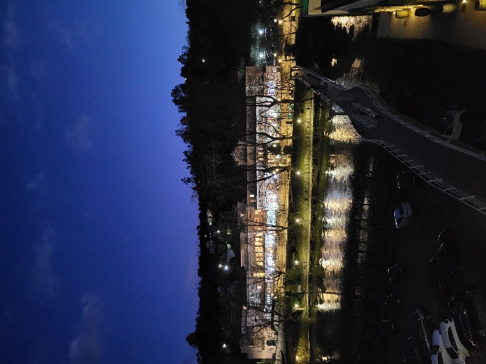 A tranquil night scene showcases a river reflecting soft lights from buildings lining its bank. Trees are silhouetted against the sky, while a bridge spans the river. Cars are faintly visible on the right side. The sky transitions from a deep blue above to darkness near the horizon. The scene evokes a peaceful, urban-meets-nature atmosphere.
