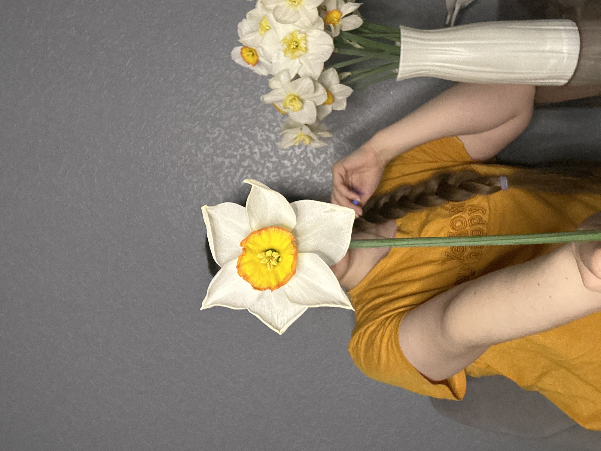 The image presents a flat lay composition on a gray surface. A single daffodil with white petals and a yellow and orange center is prominently displayed on the left. A vase containing a bouquet of white daffodils is positioned at the top right. A person wearing a mustard yellow shirt is also visible. The person's arm, hand, and braided hair are visible. The scene does not provide any information to identify the location as Saint Petersburg, Russia.