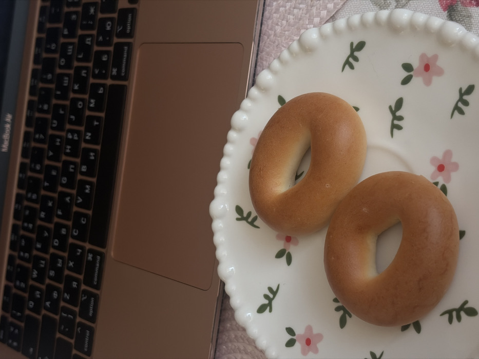The scene is an indoor setting in Saint Petersburg, Russia. The image captures a close-up perspective of two plain, golden-brown bagels resting on a white ceramic plate with a beaded rim. The plate features a decorative pattern of small pink flowers with green leaves. In the background, the top portion of a rose-gold Apple MacBook Air laptop is partially visible, displaying its black keyboard with Cyrillic and Latin characters and the trackpad area. A light pink woven material, possibly a placemat or tablecloth, is visible beneath the plate and the laptop. No individuals are present, and no active interactions or actions are depicted.