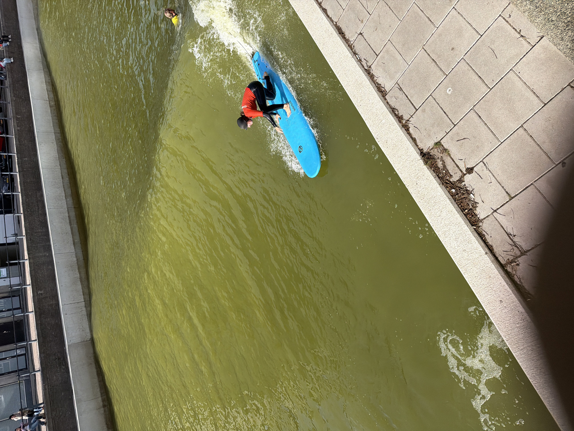 A person is surfing on a bright blue surfboard on a green, choppy body of water. They are wearing a black wetsuit, a black helmet, and an orange long-sleeved shirt. The person is in a crouched position, with their right hand extended towards the water. The surfboard has the word "VISION" printed on its side. In the background, to the right of the surfer, another person is swimming in the water, wearing a yellow shirt. The body of water appears to be a canal. The canal banks are lined with concrete walls and a paved walkway. In the far background, there is a building with windows and a railing. The scene appears to be outdoors, under daylight. The image location is in the city of Rotterdam, Netherlands.