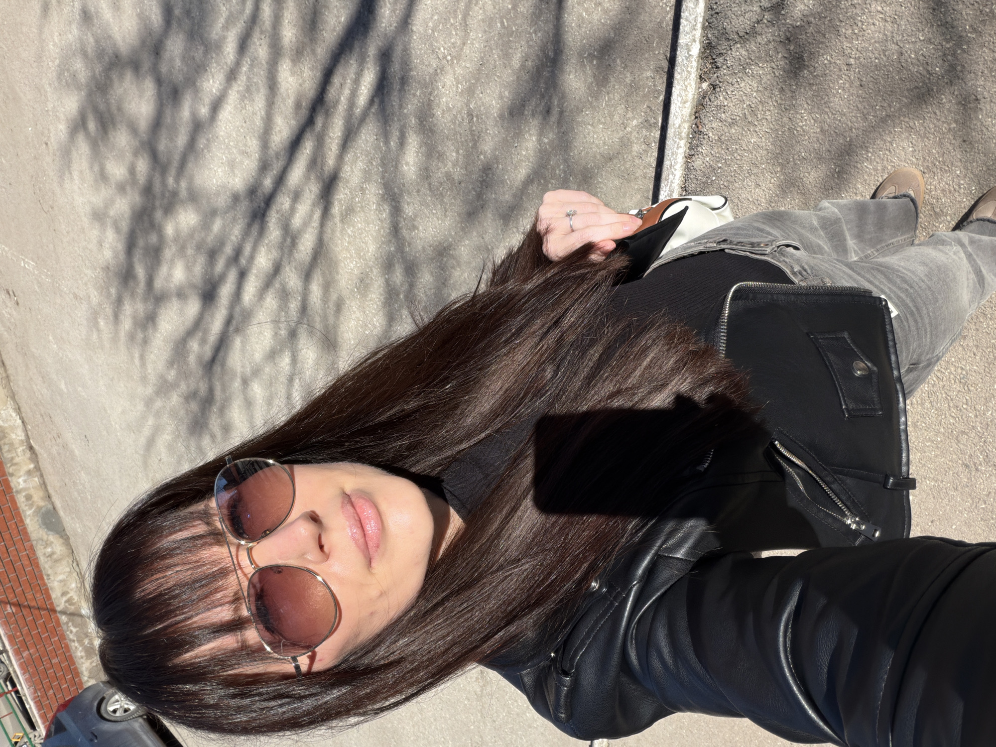A woman with long dark hair and bangs, wearing rose-tinted sunglasses, a black turtleneck, a black leather jacket, and grey jeans, is standing on a grey asphalt surface with shadows from a tree cast upon it. She is holding a white handbag with brown accents. Her left hand is visible, adorned with a ring. A portion of a brick building and a dark-colored vehicle are visible in the lower left corner of the image. The location is in the city of Perm, Russia.
