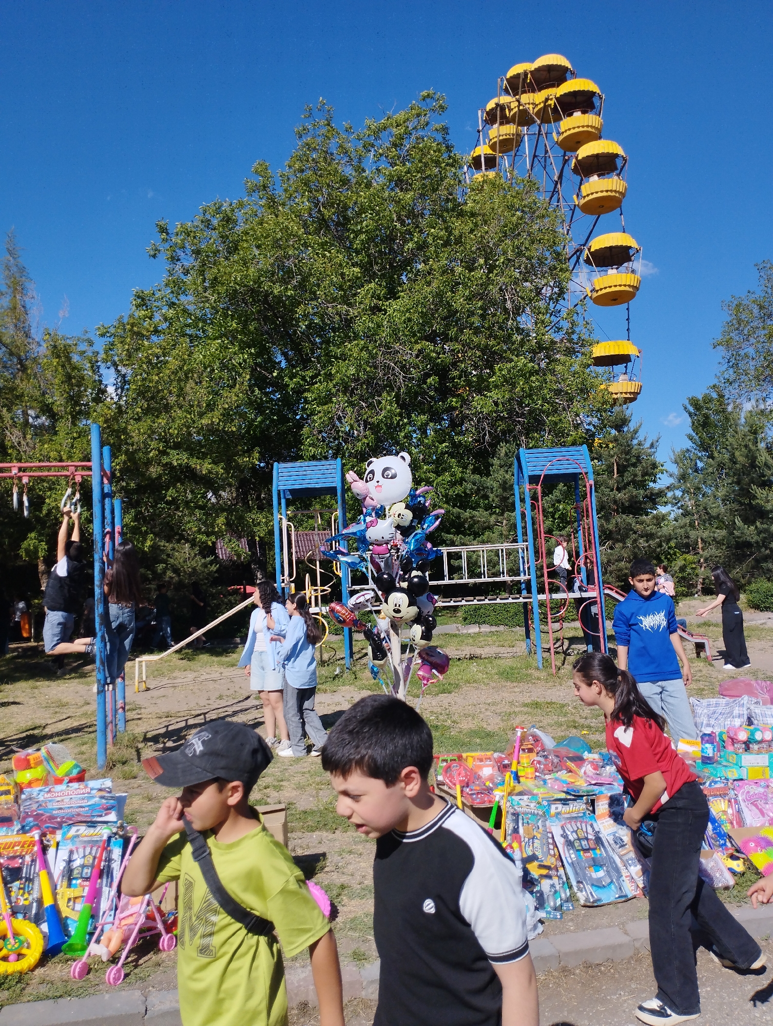 The image depicts a lively outdoor scene in Yeghegnadzor, Armenia, resembling a park festival or market on a bright, sunny day. A clear blue sky is visible above numerous green trees, which dominate the background and midground.
A prominent feature is a large, yellow-cabined Ferris wheel, partially obscured by trees, suggesting the location is adjacent to or within an amusement park. In the foreground and midground, a vibrant market stall is set up on the ground, displaying a wide array of toys and games. These include board games like "Монополия" (Monopoly), toy police sets with "Police" text visible, miniature strollers, dolls, toy vehicles, and various other children's items.
Several people, mostly children and teenagers, are scattered throughout the scene. Some are engaged in playing on blue and red metal playground equipment, such as monkey bars and climbing structures. Others are seen browsing or interacting with the merchandise. A balloon vendor is visible, holding a large bunch of character balloons, including a panda, Minnie Mouse, and Hello Kitty. The atmosphere appears casual and bustling, with people enjoying the sunny weather and the available entertainment and goods. The time of day is clearly daytime, with strong sunlight casting shadows.