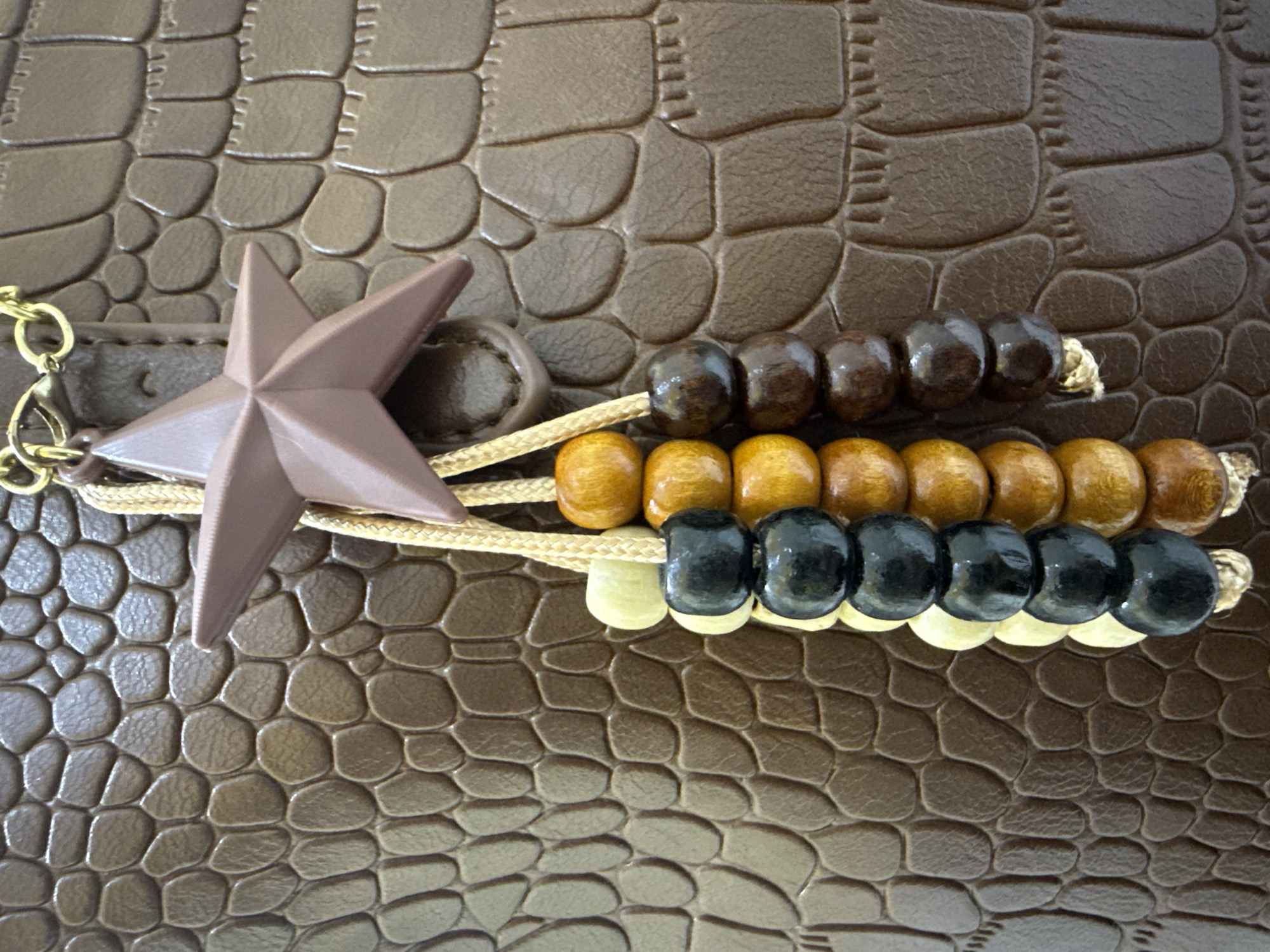 A close-up shot focuses on a brown, textured material, possibly faux leather with a crocodile-like pattern. Attached to this material is a decorative charm consisting of a dusty rose colored star pendant suspended from a gold chain. Below the star, three strings of wooden beads hang vertically. Each string features alternating beads of cream, black, and a reddish-brown or dark wood color, as well as golden-brown beads. The lighting is even, highlighting the textures and details of the objects. The background is the same textured brown material. There are no people or any contextual clues to indicate a location in Lhokseumawe, Indonesia.