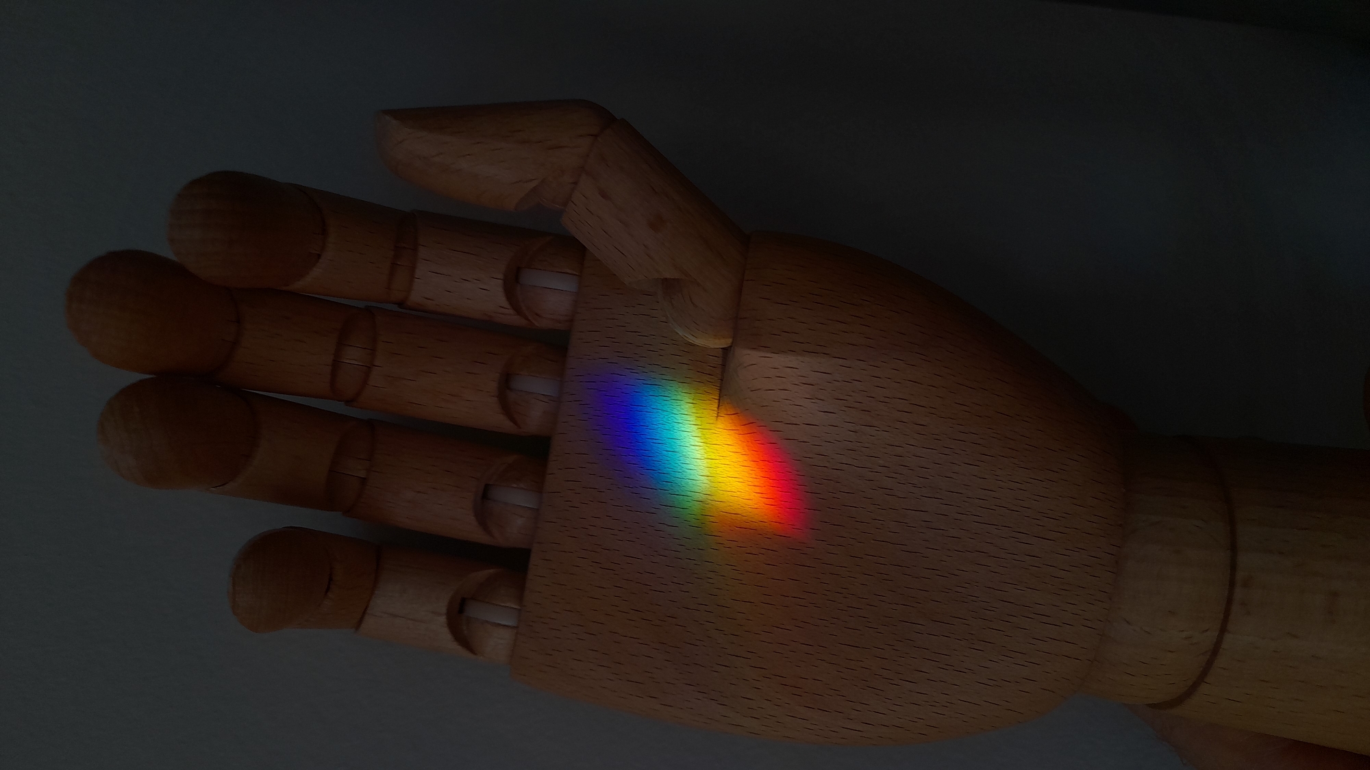 The image shows a wooden articulated hand with a rainbow reflection on its palm. The hand appears to be lying on a surface, possibly a table. The background is dark and out of focus. The colors of the rainbow are distinct, suggesting a strong light source and clear prism effect. There is no evidence in the image to determine whether the location is in the city of Saint Petersburg, Russia.