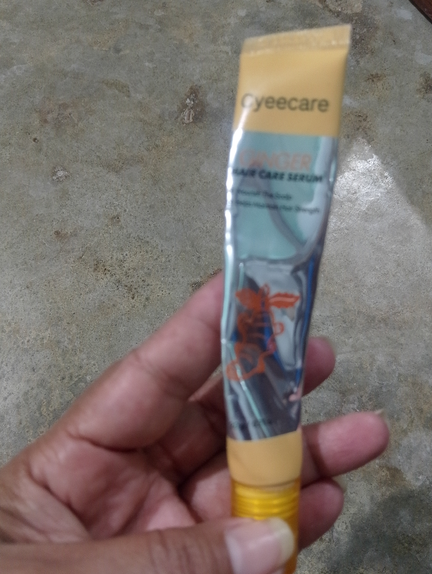 The image shows a hand holding a tube of "Gyeecare Ginger Hair Care Serum". The tube is yellow and silver with orange text and an illustration. The background is a textured, mottled gray surface, possibly concrete. The image is taken from a high angle, with the hand and tube in the foreground. No specific location in Serbelawan, Indonesia can be determined from the image.