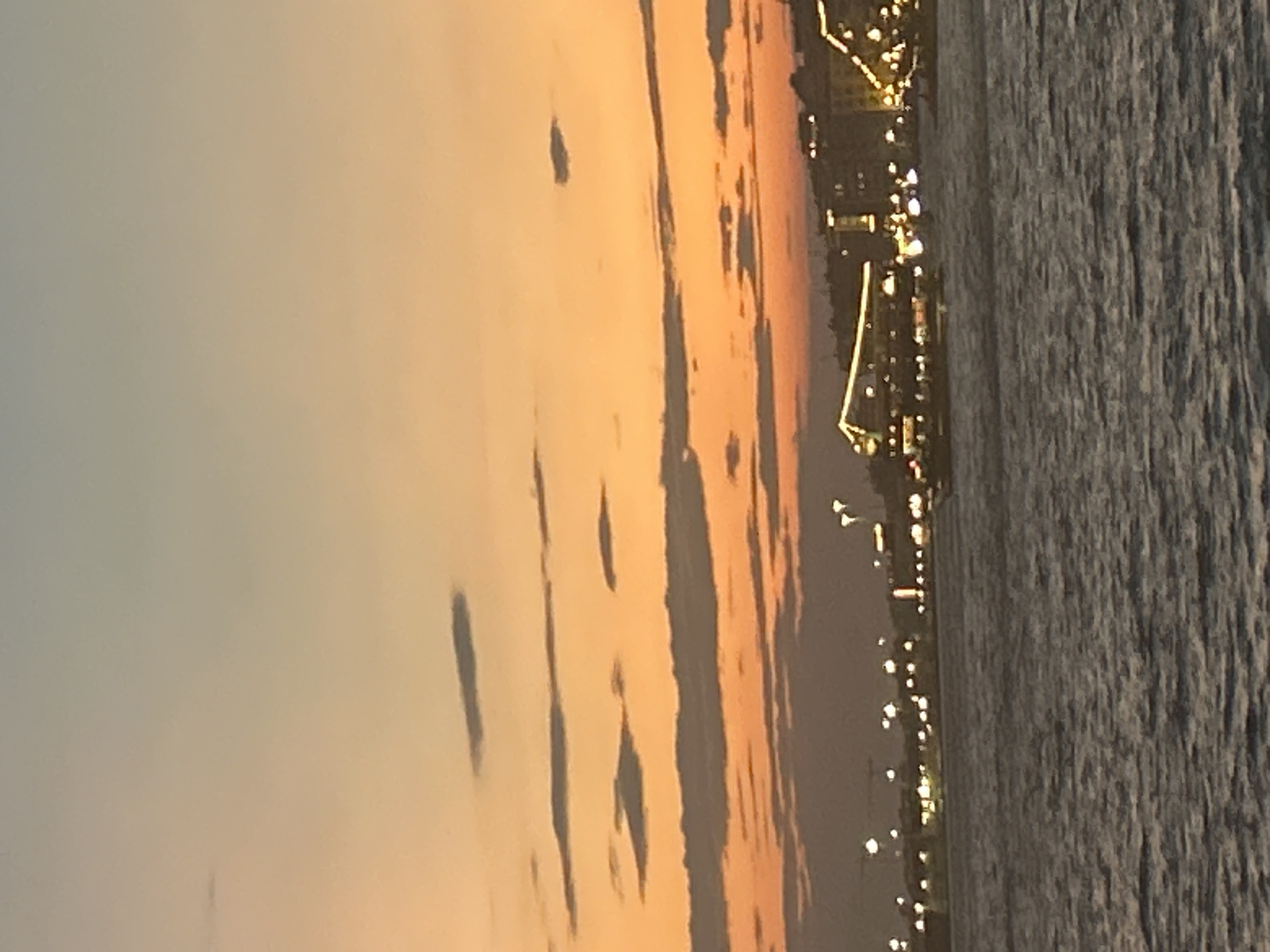 The image depicts a sunset over the Thermaic Gulf in Thessaloniki, Greece. The sky transitions from a pale blue at the top to a vibrant orange and pink hue near the horizon, with scattered clouds. In the foreground, the sea exhibits textured ripples. The background shows a dimly lit cityscape with buildings illuminated by numerous lights, suggesting it is dusk or early evening. Several boats are visible on the water, some appearing to be docked or moving. The overall atmosphere is serene and picturesque.