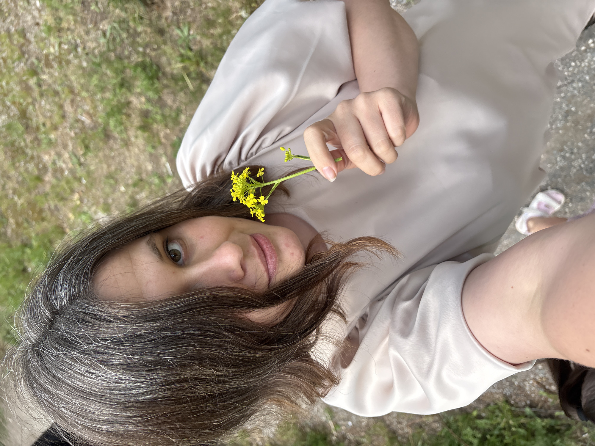 A woman is holding a small yellow flower. She has brown hair with gray roots and fair skin. She is wearing a beige, satin-like top with short sleeves. The background appears to be a mix of grass and gravel. There are no clear indicators that would definitively place the location as Chaikovsky, Russia.