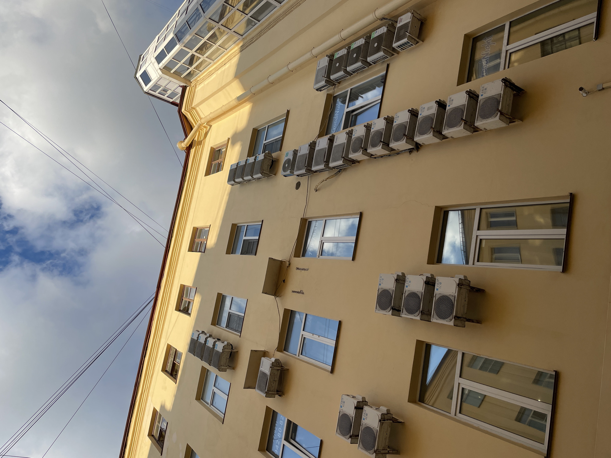 This image is a low-angle shot of a light yellow building facade in Saint Petersburg, Russia. The building has multiple windows of various sizes, and many air conditioning units are mounted on the exterior wall beneath and next to the windows. The units are white and appear to be of different models. The sky is partly cloudy, with patches of blue sky visible. The sun is shining, casting shadows on the building. There are power lines crisscrossing the sky in the upper left of the frame. A unique glass-enclosed structure juts out from the top of the building. Some of the windows have reflections of the sky and adjacent buildings. One window has the text "GoodNight" visible on it. Another window has a phone number with Cyrillic text visible. The overall impression is of an urban environment, possibly residential or office buildings, where air conditioning is widely used. The time of day appears to be daytime, and the weather is fair.