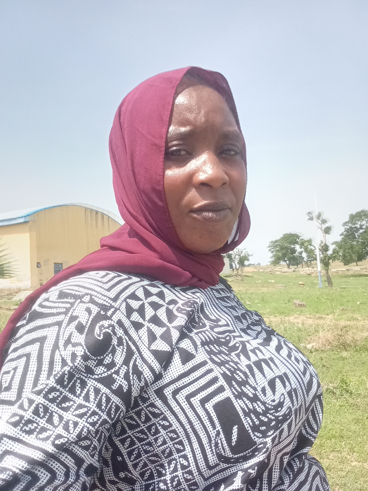 A close-up, slightly high-angle portrait shows a woman outdoors under a bright, clear sky. She has dark skin and is looking directly at the camera with a neutral to serious expression. Her face appears somewhat shiny, possibly from sweat or bright light. She is wearing a maroon-colored hijab that covers her hair and neck, and a black and white patterned top with intricate geometric and abstract designs.
The background features a rural or semi-urban landscape under bright daylight. To the left, a light yellow or beige building with a light blue or gray arched roof is visible. The building shows some signs of wear and dirt streaks. Below the building's window, faint text "KILL 14" is visible. To the right and behind the woman, a vast expanse of dry, yellowish-green grassy land is present, with several scattered trees and some bare branches. A tall, thin light-colored pole is also visible in the distance. The overall setting suggests a sunny day in an open, possibly agricultural or undeveloped area. The angle of the shot suggests a selfie.