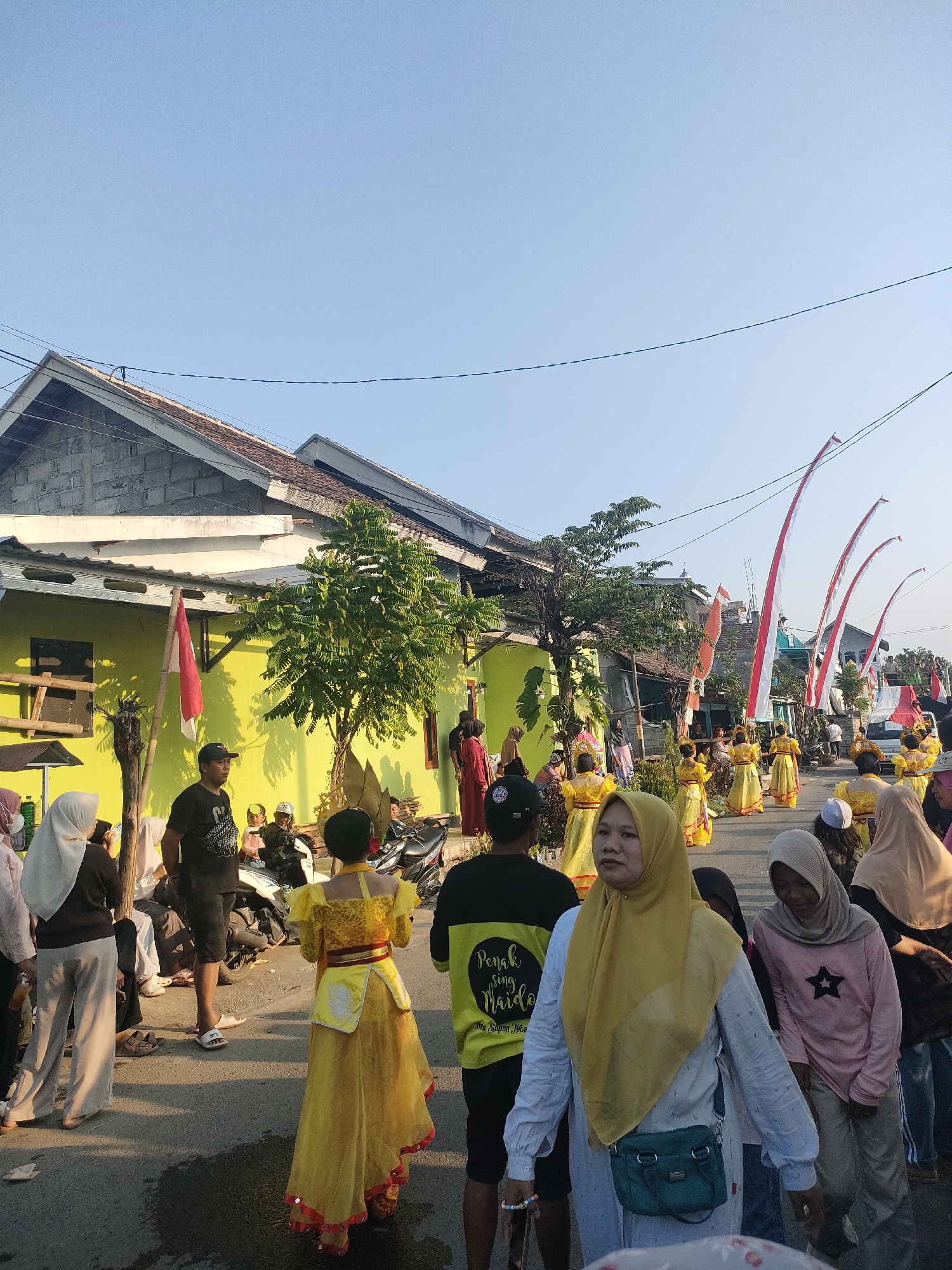 In Tongaswetan, Indonesia, on a street lined with houses and adorned with Indonesian flags, a procession is underway. Several individuals, primarily women and children, are dressed in traditional yellow attire and are participating in what appears to be a cultural performance or parade. Some are walking in unison, while others observe from the sidelines. The street is paved, and a few motorcycles are visible. The atmosphere is festive, indicated by the decorations and the gathering of people. The sky is clear and blue, suggesting a sunny day.