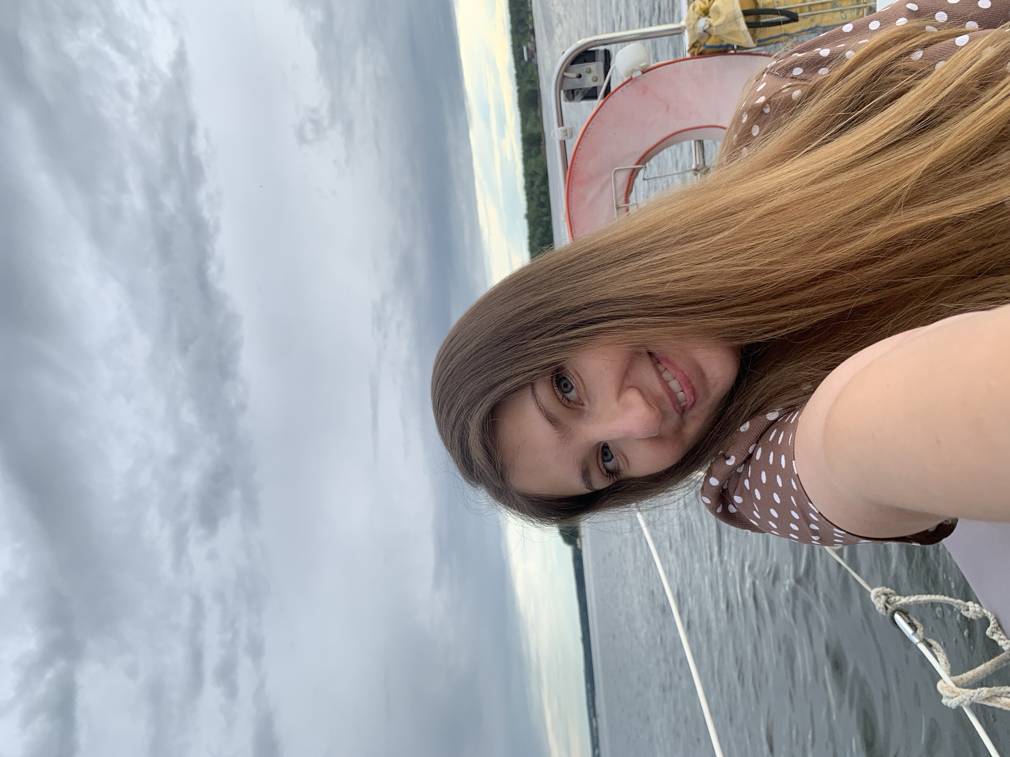 A young woman with long, light brown hair and blue eyes takes a selfie on a boat. She is smiling and wearing a brown dress with white polka dots. In the background, the water is calm, and the sky is cloudy. A life preserver is visible on the boat. The location is likely on the Kama River in Chaikovsky, Russia.