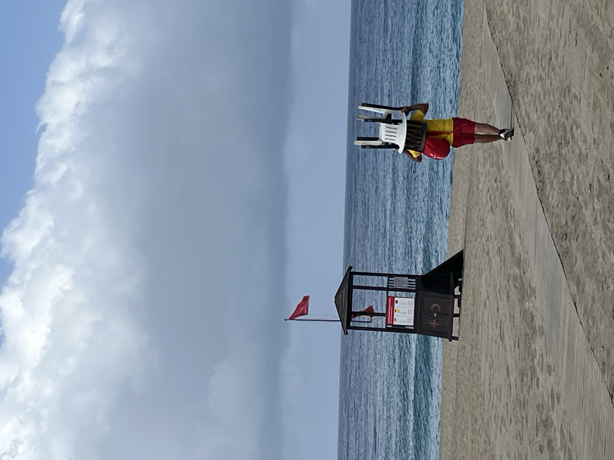 A lifeguard carries chairs on the beach in Alanya, Turkey. The sky is cloudy and blue. The lifeguard wears a yellow shirt and red shorts, and the ocean extends behind him. A lifeguard tower stands on the beach with a red flag flying. The sand is light in color.