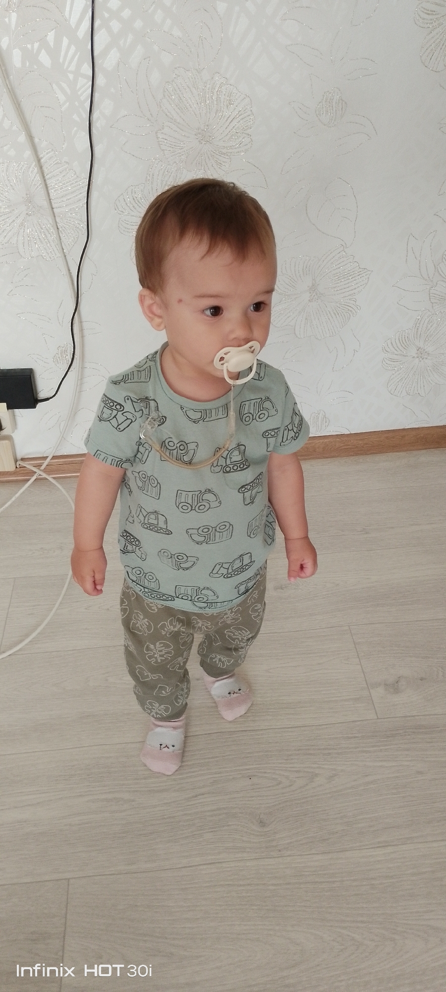 A full-body, eye-level shot shows a young Caucasian child, appearing to be a toddler, standing on a light-colored wooden floor. The child has light brown hair and is wearing a light blue t-shirt with a repeating pattern of various construction vehicles, and khaki-colored pants with a leaf pattern. The child also has on light pink socks with a white animal face design on the front. A white pacifier is in the child's mouth, attached by a clear and beige string to the t-shirt. The child is looking off to the right of the frame. Behind the child is a white wall with a textured floral pattern. A black electrical cord hangs vertically on the left side of the wall, connected to a black electrical adapter. The environment suggests an indoor setting, possibly a home. There is no indication of the child's specific location within the city of Kedrovy, Russia.