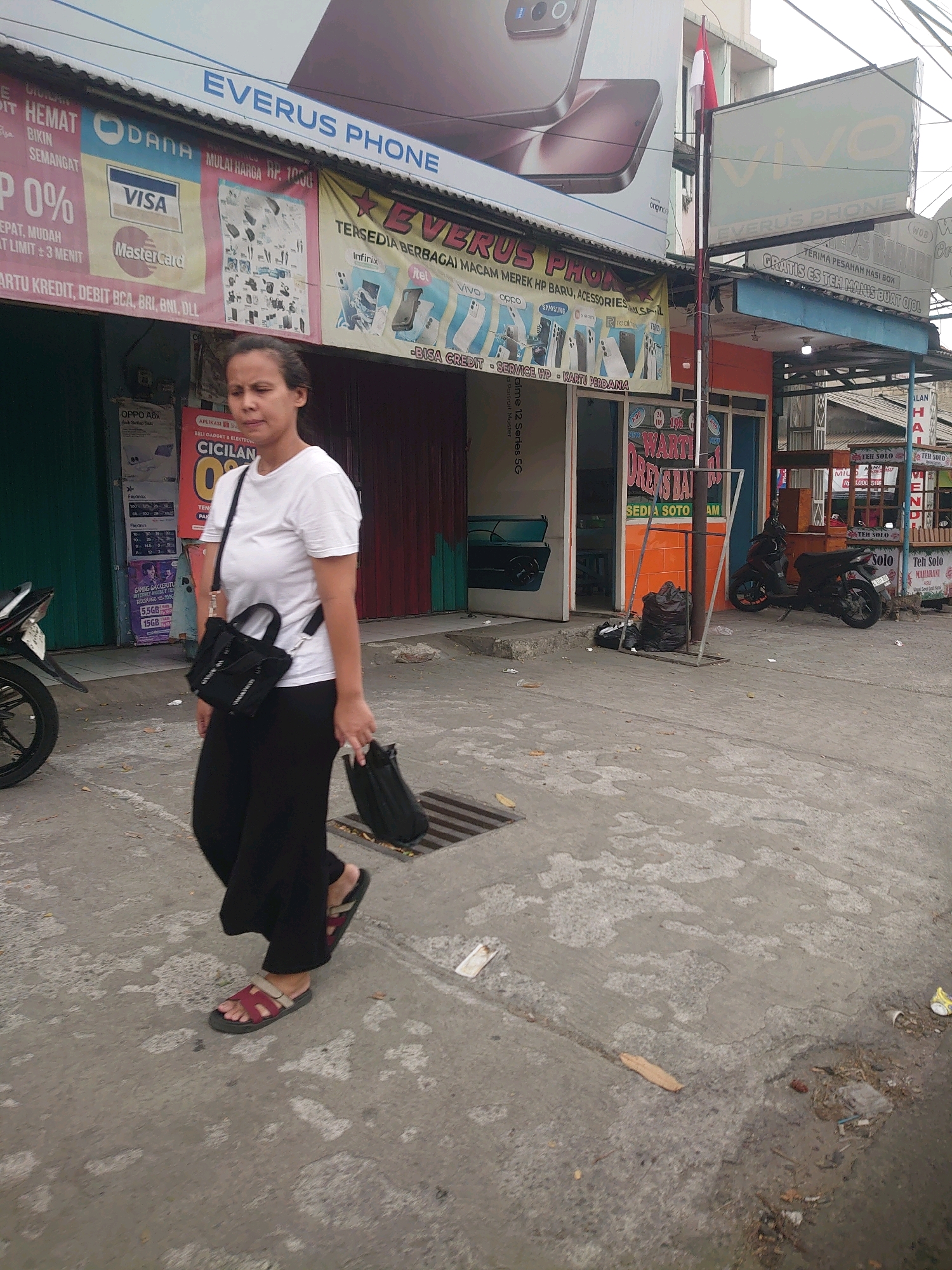 A woman walks on a sidewalk in front of a street-level storefront in Depok, Indonesia. She is wearing a white t-shirt, black pants, and red and black sandals. She carries a black tote bag over her right shoulder and a black bag in her right hand. The storefront features signage for "EVERUS PHONE" and advertisements for mobile phones and payment services. A motorcycle is parked on the street, and trash bags are visible near a metal pole. The sidewalk is made of concrete and appears wet in places, with debris scattered around.