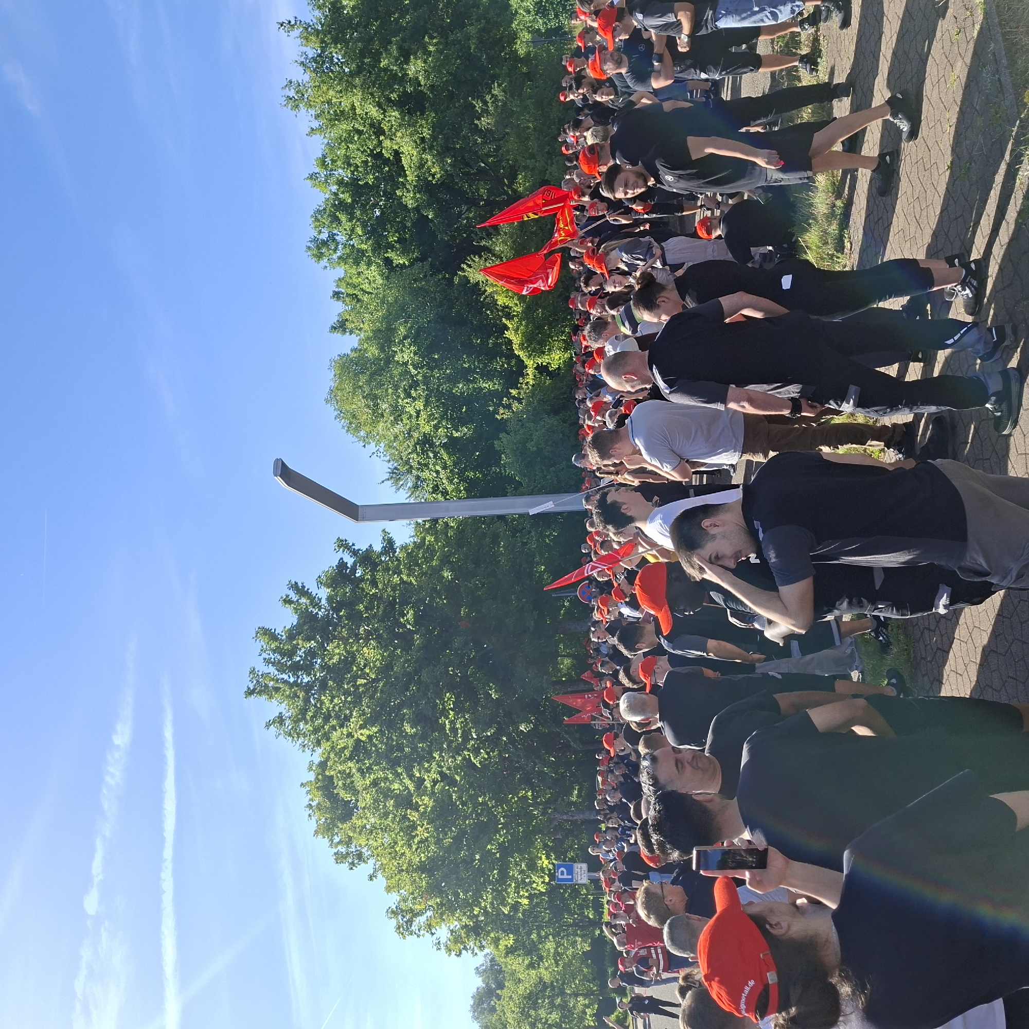 The image shows a large group of people gathered outdoors, likely for a protest or demonstration. Many individuals are wearing red caps and some are holding red flags. The environment appears to be a paved area with grass and trees in the background. A tall, modern lamp post is visible in the center of the image, and a blue parking sign is present to the left. The sky is clear and blue with some thin white clouds. The people are diverse in age and gender, and their interactions suggest a shared purpose or event. Some individuals are holding up their phones, possibly to record or take pictures. The overall atmosphere seems to be one of collective engagement. The presence of flags and the number of people suggest a significant gathering. The context of Rastatt, Germany, implies a specific regional event or demonstration.