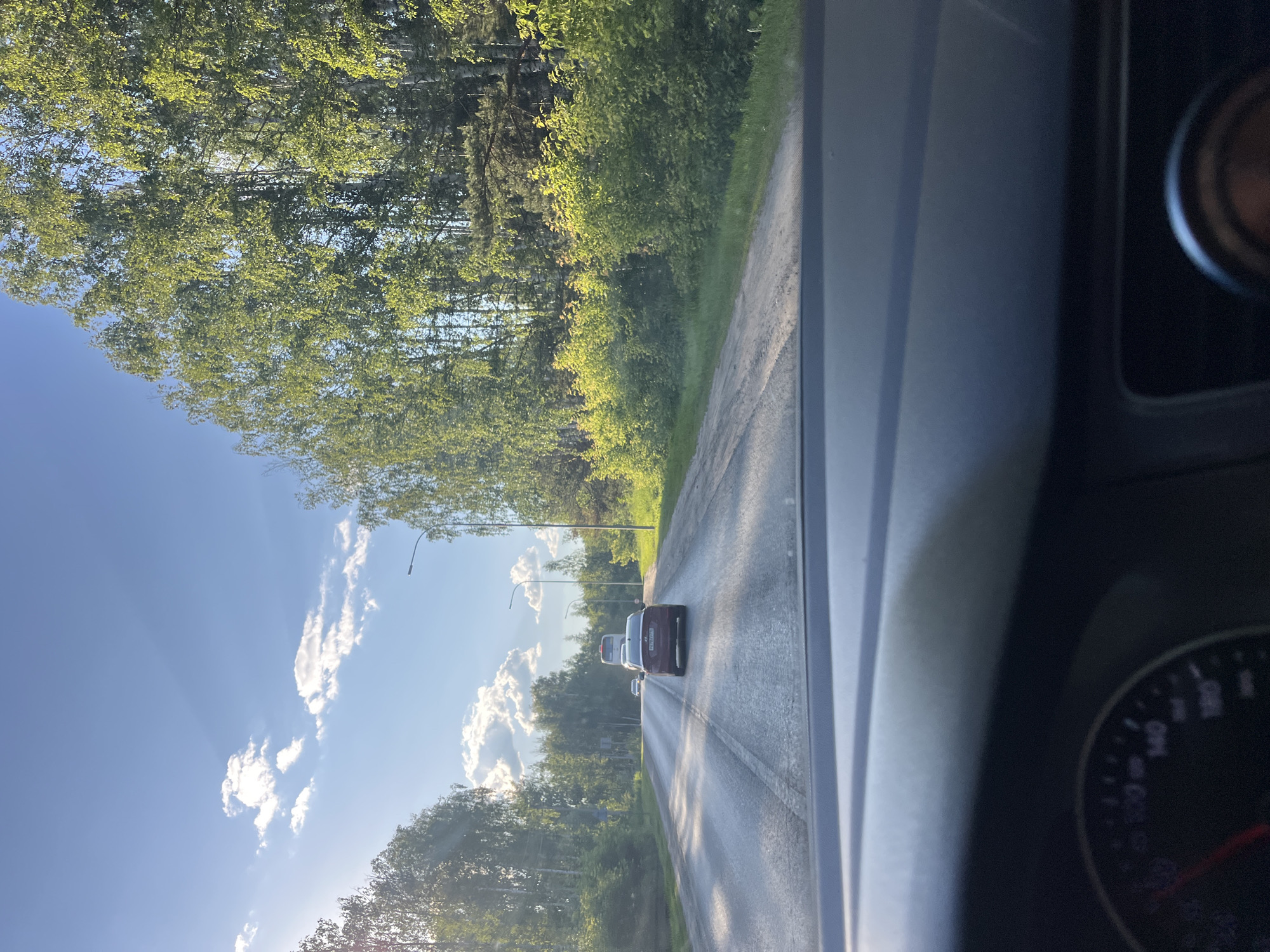 A forward-facing view from inside a vehicle shows a paved road with light traffic under a blue sky with scattered clouds. The road is wet and reflects the sunlight. The right side of the road is bordered by a dense line of green trees. A red car is visible in the foreground, driving in the same direction as the camera. Further down the road, a white bus and another car are also visible. The dashboard of the vehicle, including a speedometer, is partially visible in the lower portion of the frame. The scene appears to be in a rural or suburban area, consistent with Vasilyevo, Russia, given the presence of birch trees and the general landscape.