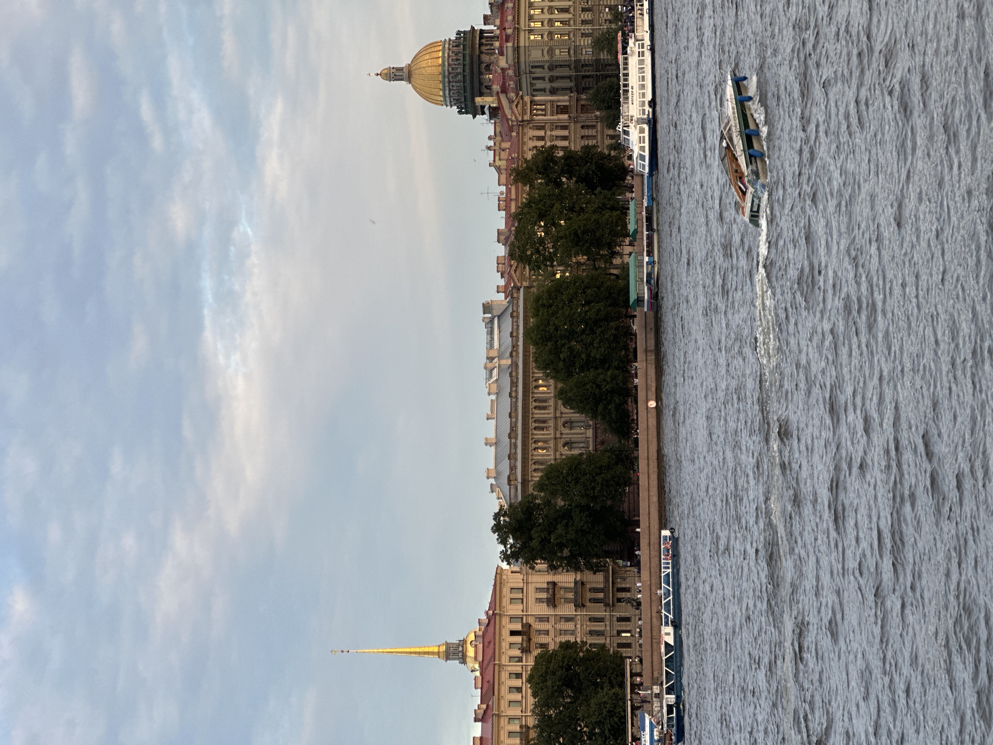 The image depicts a view of the Neva River in Saint Petersburg, Russia, with the cityscape lining the embankment. On the left, the spire of the Peter and Paul Fortress pierces the sky, distinguished by its golden color. Adjacent to it are historic buildings with red-tiled roofs. Further to the right, the golden dome of St. Isaac's Cathedral dominates the skyline. Several boats are visible on the river. Two large tour boats are docked near the embankment, with people on board. In the foreground, a smaller motorboat speeds across the water, leaving a wake. Two individuals are seated in the motorboat. The sky is overcast with a mix of light and dark clouds, suggesting it is either early morning or late evening. The water of the Neva River is choppy, with visible ripples and waves. The overall scene captures a characteristic view of Saint Petersburg's architectural grandeur and maritime activity.