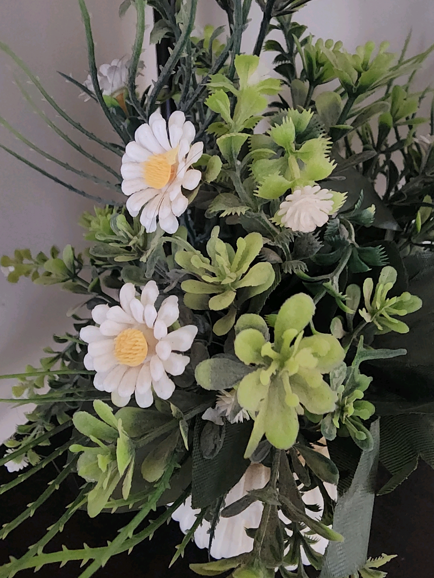 A close-up, eye-level, slightly angled shot displays an artificial floral arrangement. The arrangement features white daisy-like flowers with yellow centers, interspersed with various shades of green artificial foliage. Some of the green leaves are plump and rounded, while others are elongated and spiky. The background is a plain, neutral-colored wall. The lighting is soft, casting gentle shadows and highlighting the textures of the artificial plants. No people are visible in the image. The composition focuses on the details of the floral arrangement, with a shallow depth of field blurring the background.
