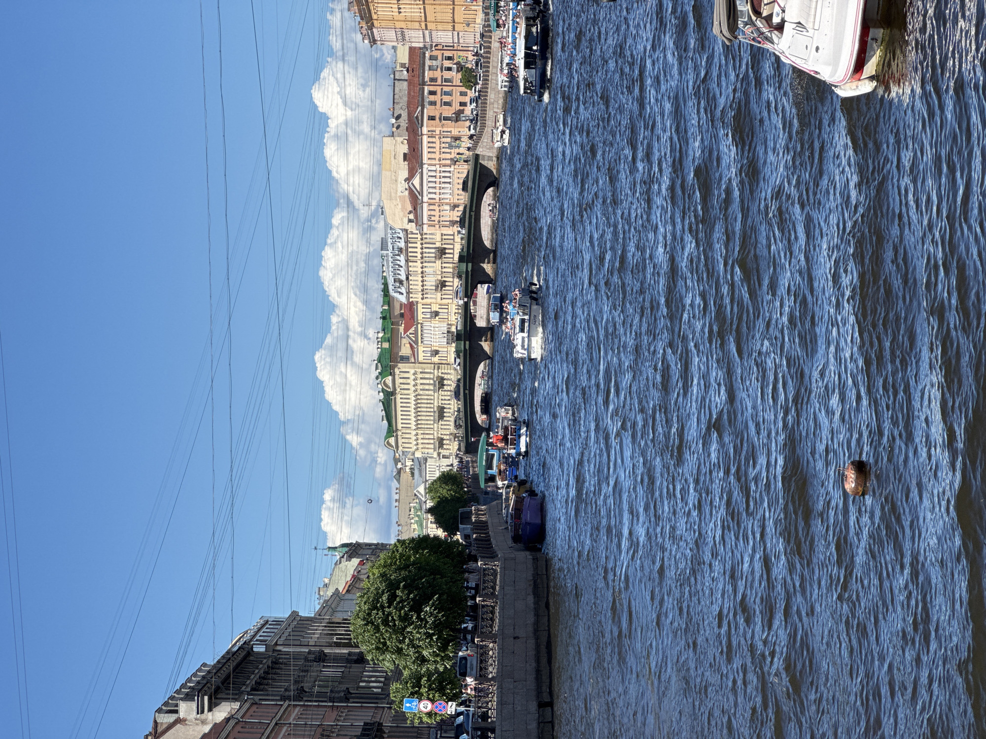 This image captures a vibrant daytime scene along the Fontanka River in Saint Petersburg, Russia.
The foreground is dominated by the dark blue, rippled water of the river. A reddish-brown, cylindrical buoy or floating object is positioned in the lower-center water. In the lower right, the stern and partial seating area of a white recreational boat with red trim and a dark canopy are visible.
Along the left bank, a stone embankment features a dark metal railing. Above the embankment, a pedestrian walkway is bordered by trees and a road with parked cars and moving traffic. Several pedestrians are visible. A blue road sign on the left indicates a bicycle path and a 40 km/h speed limit. Behind this, multi-story historic buildings with varied architectural styles line the street. Several smaller boats are docked along this bank.
The river itself is populated with various vessels. Several tour boats, laden with passengers, are in motion. One prominent white tour boat displays the text "ГАРИ ШОУ" (GARI SHOU) on its side. Another dark tour boat on the right explicitly identifies its location with "ФОНТАНКА" (FONTANKA) written on its hull.
In the middle distance, a multi-arched stone bridge spans the river, accommodating vehicular and pedestrian traffic. Beyond the bridge, along the right bank, a continuous row of multi-story historic buildings with diverse facades in yellow, orange, and beige tones, some featuring green roofs, extends into the background. People and vehicles are discernible on this distant embankment.
The sky above is bright blue with scattered white cumulus clouds, and multiple dark power lines or communication cables crisscross the upper portion of the frame.