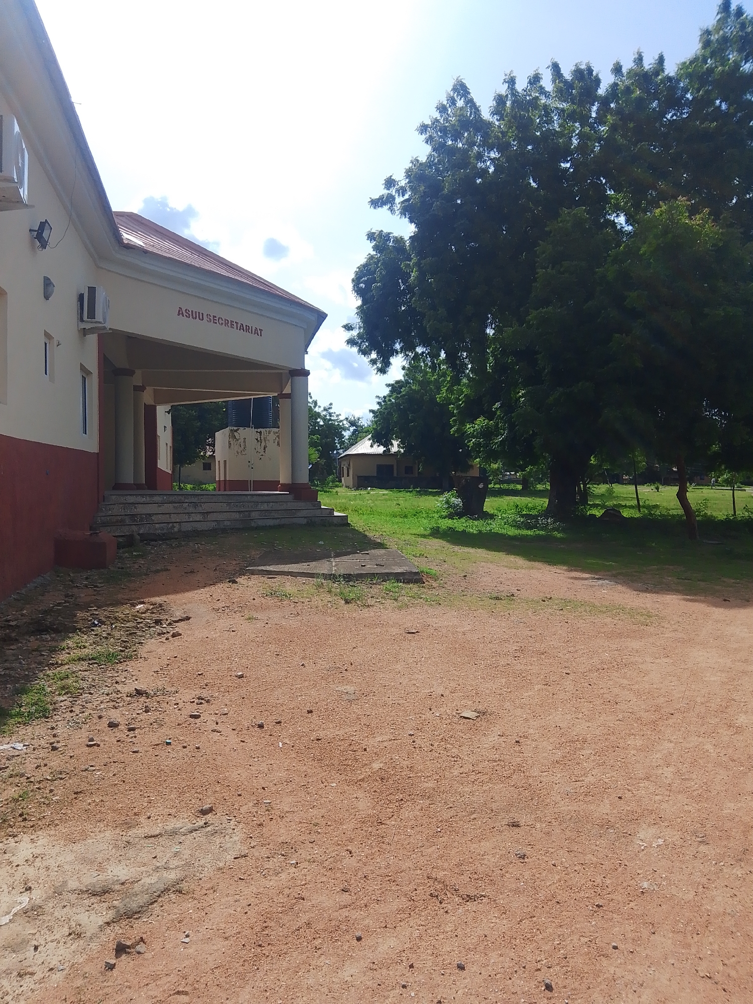 This media file captures an exterior view of a building labeled "ASUU SECRETARIAT". The building has a cream-colored facade with a red lower section. It features an overhang supported by columns, with a set of concrete stairs leading up to it. Two air conditioning units are visible on the exterior wall.
The scene is outdoors, with lush green grass and several large, mature trees in the background. Another building with a light-colored exterior and a red roof is visible further back in the grassy area. The ground in the foreground is a reddish-brown, dirt or gravel pathway, with scattered small stones and debris.
The sky is bright blue with some white clouds, suggesting it is daytime, likely late morning or early afternoon, given the lighting. The weather appears to be clear and sunny. There is no discernible activity or people in the image.
The visible text "ASUU SECRETARIAT" is prominently displayed on the building, indicating it is likely a meeting or administrative center for the Academic Staff Union of Universities. The overall impression is one of a somewhat rustic, academic or administrative environment in a tropical or sub-tropical location, possibly within a university campus or similar institution in Jalingo, Nigeria.