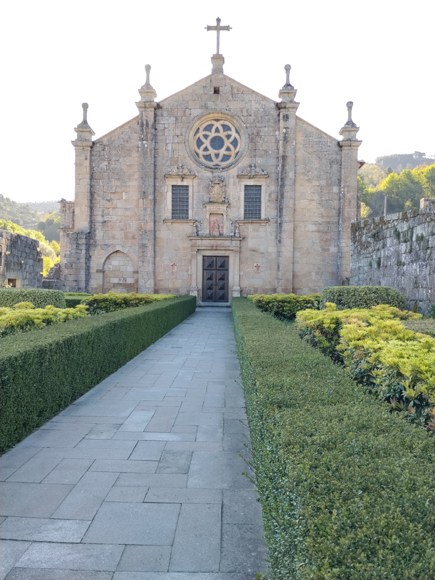 The image depicts the exterior of a stone building, likely a church or monastery, located in Tarouca, Portugal. A central stone pathway leads to the building's entrance, bordered by meticulously trimmed green hedges and patches of yellow-green foliage. The building features a symmetrical facade with a rose window above two rectangular windows. A cross adorns the apex of the roof. To the right of the building is a stone wall extending away from the main structure. The surrounding environment consists of rolling hills covered in trees. The scene appears to be captured during daylight, with soft sunlight illuminating the stone facade.