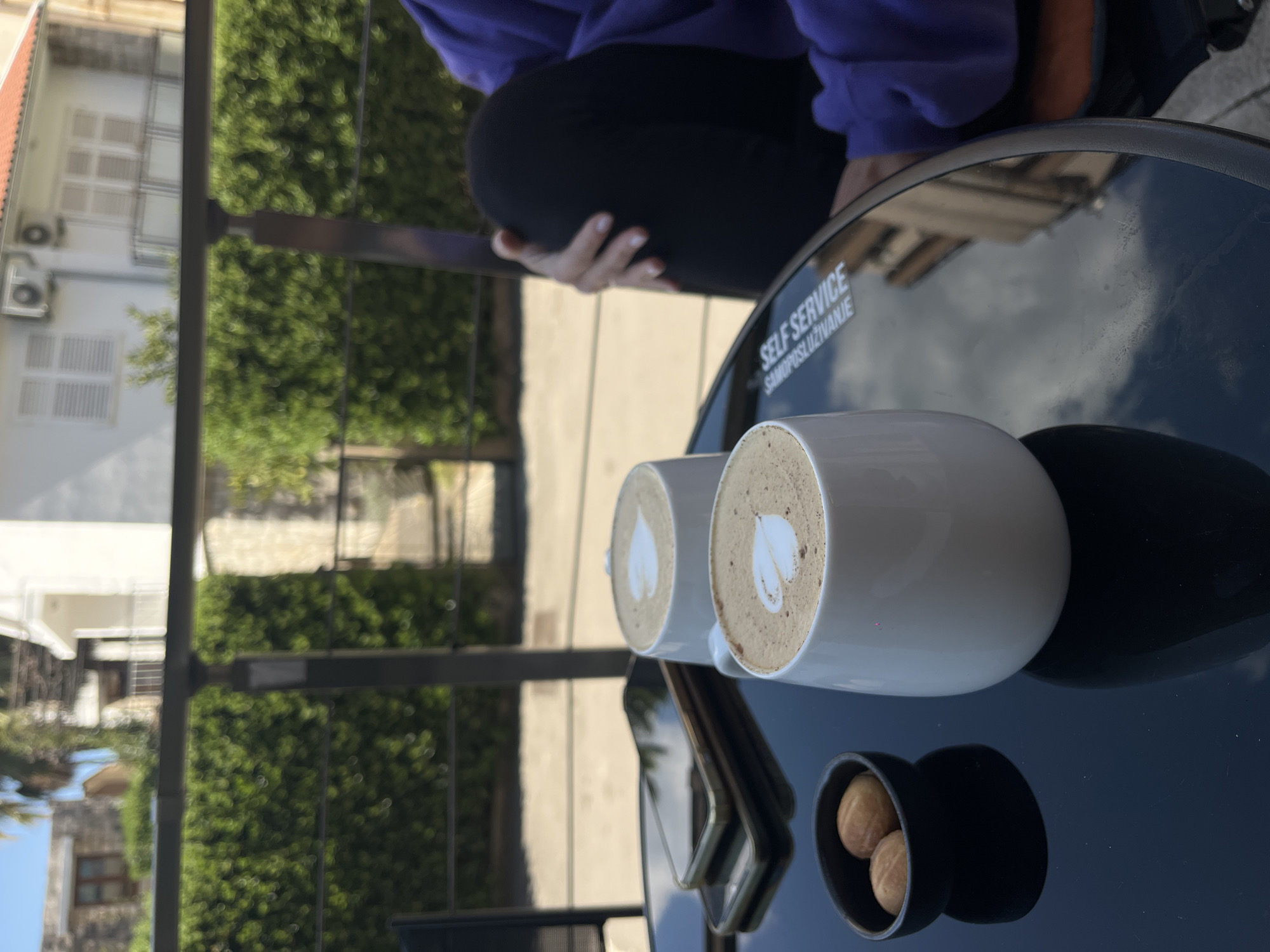 The scene is an outdoor setting, likely a café in Budva, Montenegro. Two white mugs containing lattes with latte art are placed on a black, reflective table, alongside a small black dish containing three pastries. A person wearing a purple top and black pants is sitting near the table. A cell phone rests on the table. The sticker on the table has the text "SELF SERVICE SAMOSLUŽIVANJE". Buildings with white facades and green trees are visible in the background.