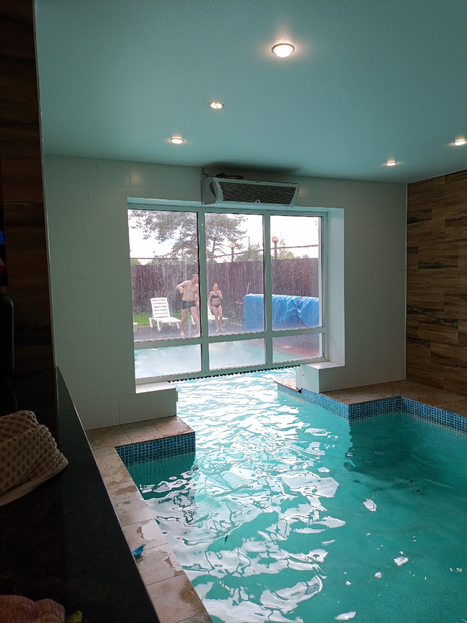 The image depicts an indoor swimming pool area in Chaikovsky, Russia, offering a view of an outdoor space through a large, multi-paned window. The indoor pool, filled with turquoise water, occupies the foreground and mid-ground, with its surface showing ripples and reflections of light. The pool's edge is tiled with small blue and white mosaic patterns, complementing the light-colored floor tiles surrounding it.
The interior features a light-colored ceiling equipped with several recessed spotlights. A wall-mounted heating or air conditioning unit is positioned above the main window. The room's walls are clad in white tiles, contrasting with a section of warm-toned wooden paneling on the right. In the immediate left foreground, a dark counter or surface holds a textured, light-colored item, possibly a folded towel.
Beyond the window, two people, a man and a woman, are visible standing outdoors. The man wears dark swim trunks, and the woman is dressed in a dark bikini. They are situated near two white plastic lounge chairs. Streaks of water are clearly visible on the window glass, suggesting ongoing rain or a strong water spray outside. The background beyond the individuals includes a dark wooden fence, some trees, and a large blue tarp covering an object. The lighting indicates a daytime setting, likely overcast due to the soft, diffused light. No text is visible in the image.