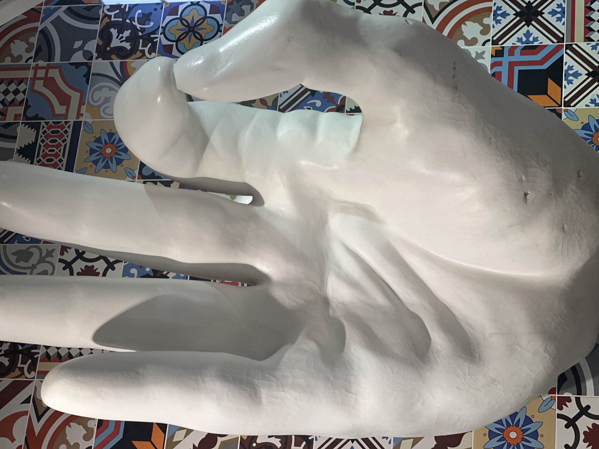 A close-up shot depicts a large, white, glossy sculpture of a human hand against a backdrop of colorful, patterned tiles. The hand is oriented palm-up, with the fingers slightly curled inwards. The tiles behind the hand exhibit a variety of geometric and floral patterns, creating a vibrant and textured background. The scene appears to be indoors, likely within a building or gallery space. The exact location in Kazan, Russia is unknown.