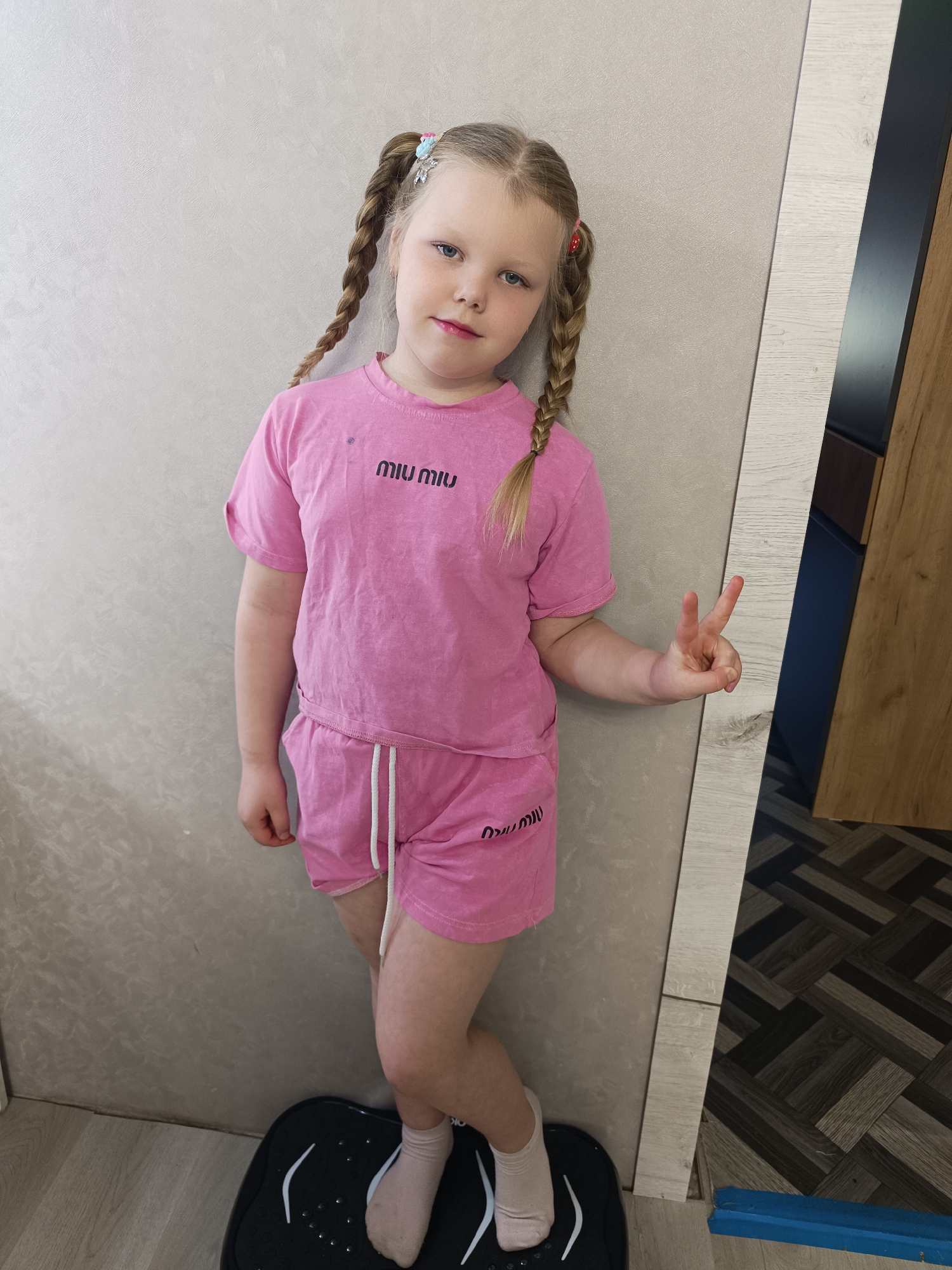 A young girl is standing indoors, posing with a peace sign. She has blonde hair styled in braids with colored accessories, and she is wearing a pink matching top and shorts set. The outfit has the words "MIU MIU" printed on both the shirt and the shorts. She is wearing light-colored socks. The girl is standing on a black electronic device of some kind. She is leaning against a light-colored wall near a doorway. The floor has a tiled pattern in dark shades.