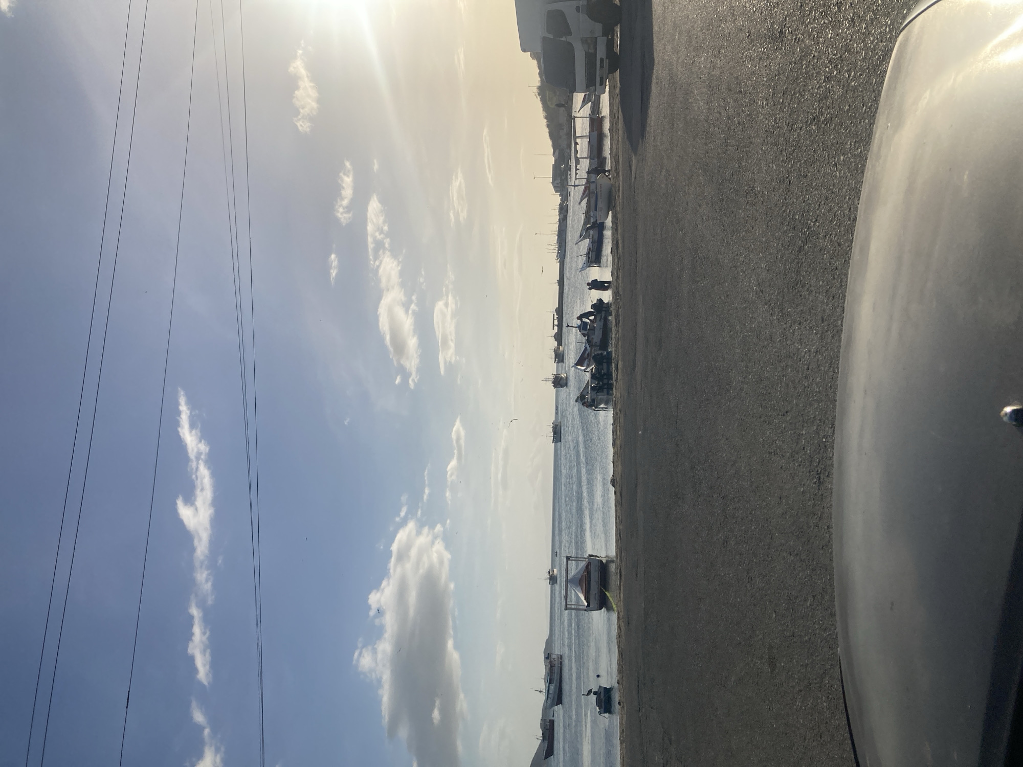 The image depicts a coastal scene in Juan Griego, Venezuela, at sunset. In the foreground, the hood of a vehicle is visible, suggesting the photo was taken from a car. The middle ground features a paved area leading to a body of water where several boats are moored. Some of these boats appear to be small fishing vessels, while others are larger. A white truck is parked on the right side of the paved area, near some of the boats. The water itself is calm, reflecting the sky, and a few birds are visible in flight. In the background, the coastline is lined with buildings and vegetation. The sky is a mix of blue and white clouds, with the sun setting on the right, casting a warm glow across the scene. There are also power lines stretching across the upper portion of the image. Several people are visible on the shore, interacting with the boats, possibly engaged in loading or unloading activities.