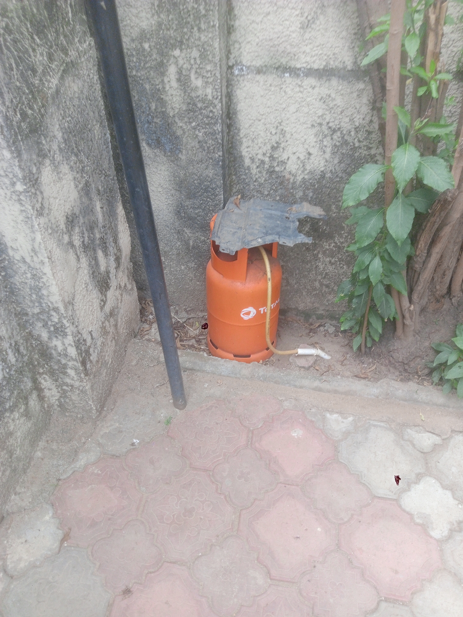 The image shows an orange Total gas cylinder in an outdoor setting in Kubwa, Nigeria. The cylinder is positioned near a wall and a black pole, with some foliage and trees visible in the background. A section of black plastic is placed on top of the gas cylinder. The cylinder is connected to a hose with a white nozzle. The ground is paved with patterned tiles. The overall environment suggests an informal or residential area. There are no people visible in the frame.